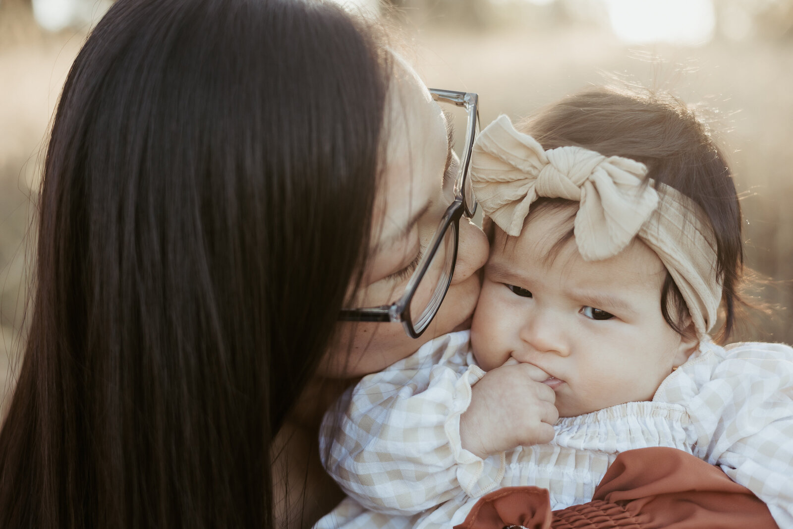 Baby girl wearing her headband and has fingers in her mouth while mum  kisses the side of her head.