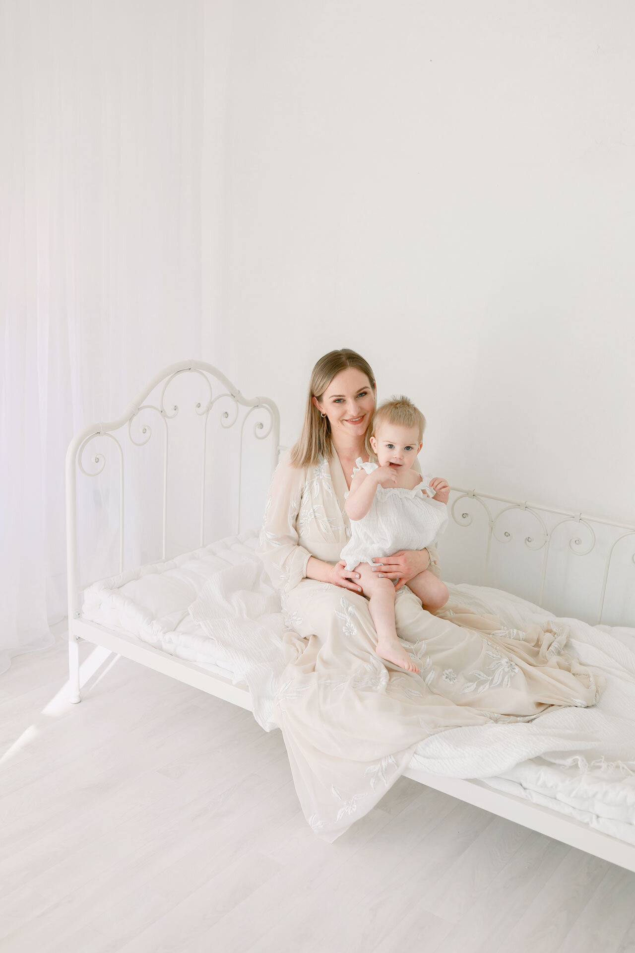 Babyshooting bei Babyfotografin im Fotostudio Bielefeld. Auf weißem Bett sitzt eine glückliche Mama in langem weißen Kleid mit Baby auf dem Schoß.