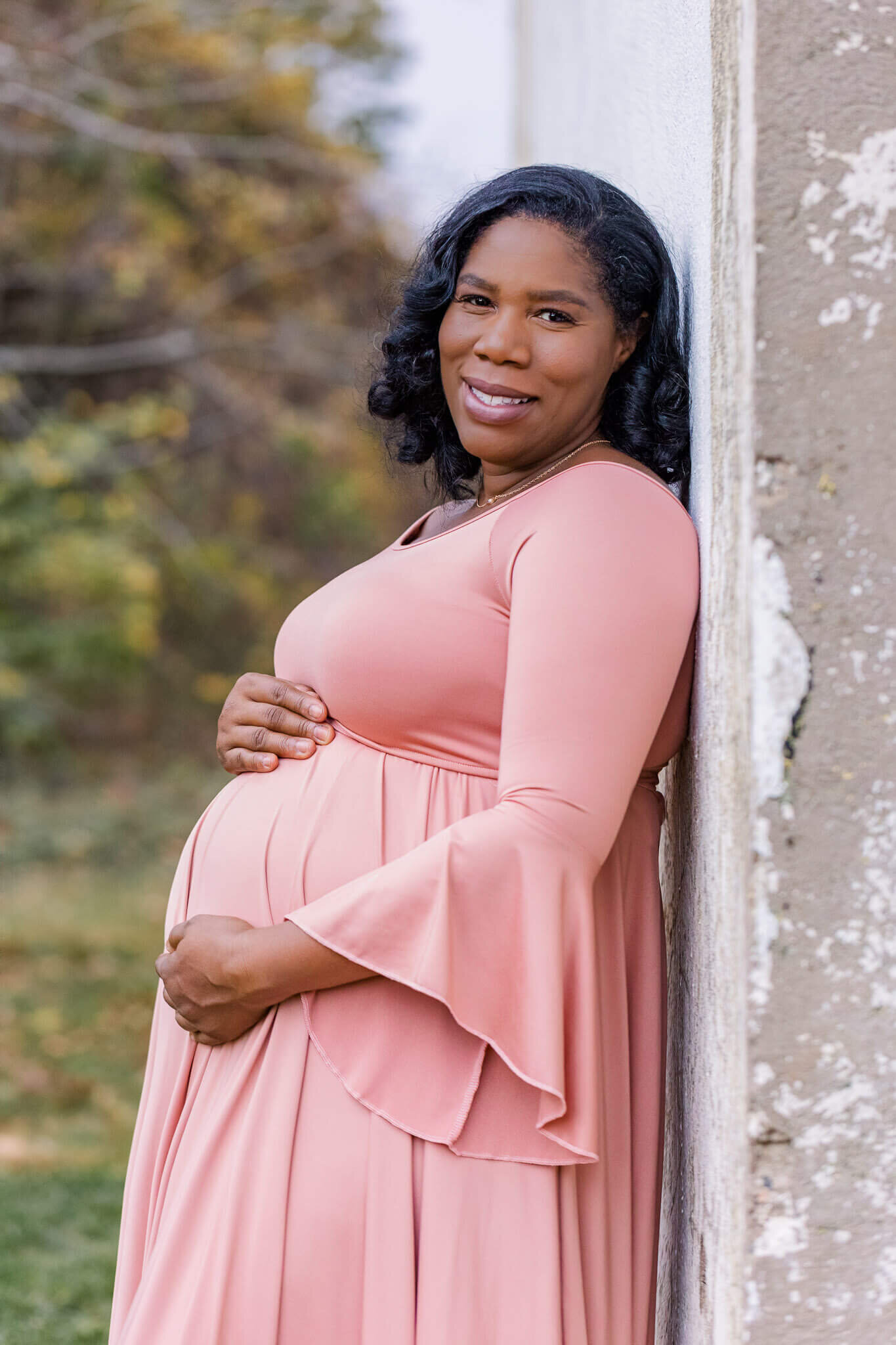 A pregnant woman wearing a pink dress and leaning against a building at Fort Hunt Park in Alexandria.