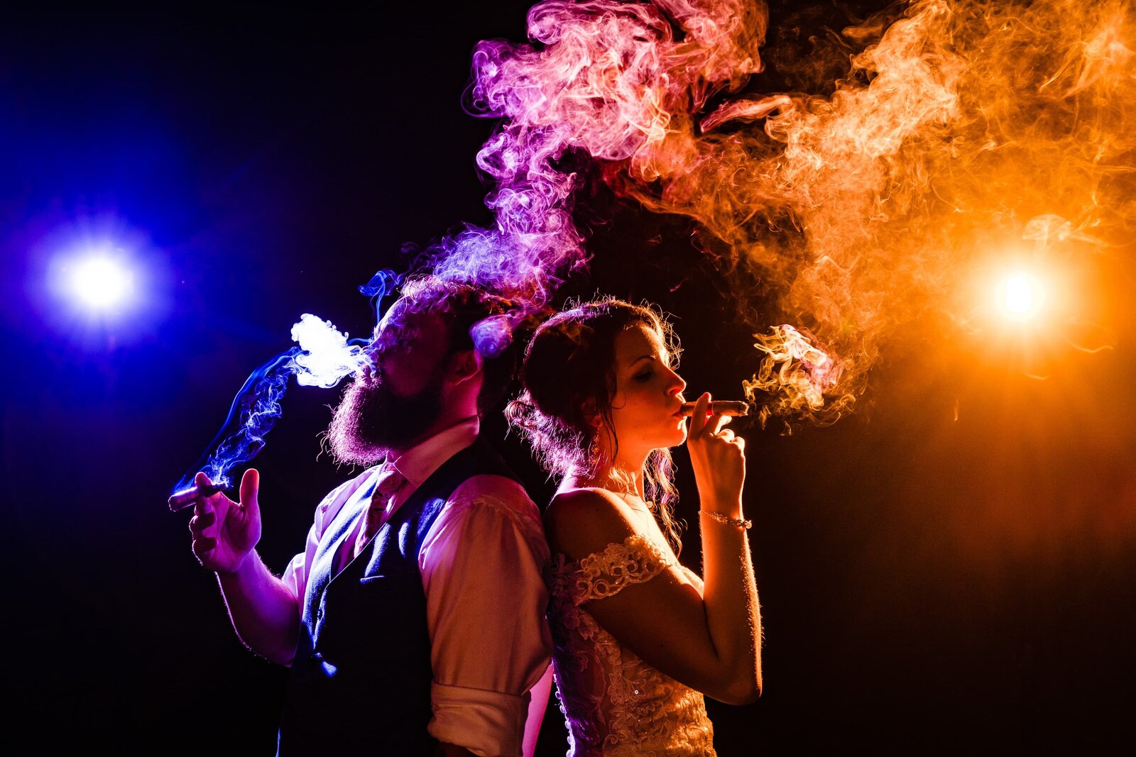 Couple dressed in wedding attire stand back to back while smoking cigars. The cigar smoke is creatively backlit with orange and purple lights.