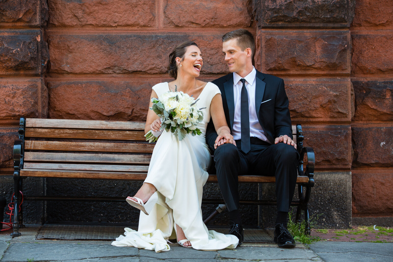 couple laughs on bench in front of brick wall