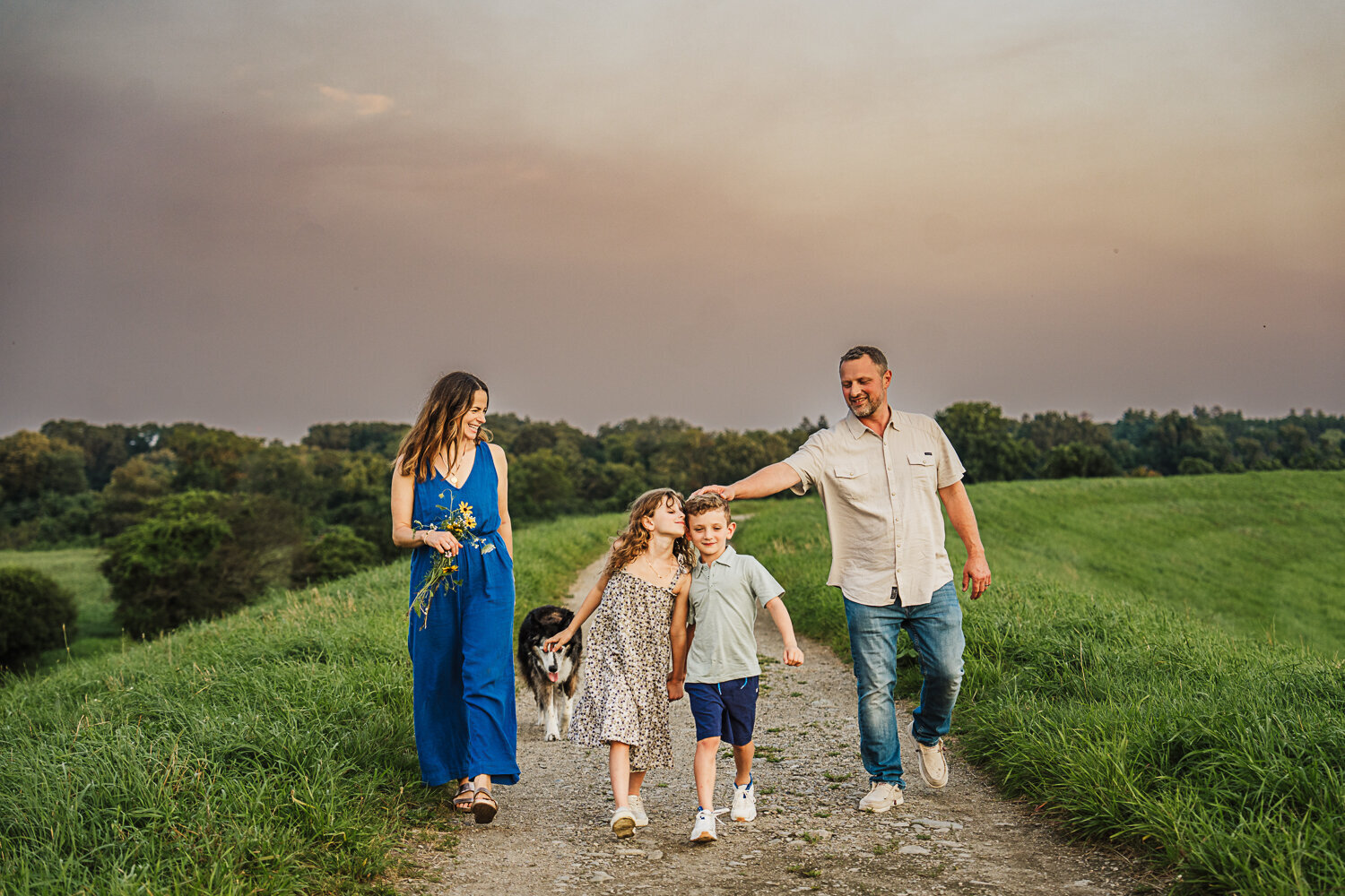 family with elementary aged kids and elderly dog walk together on a path through a field at sunset