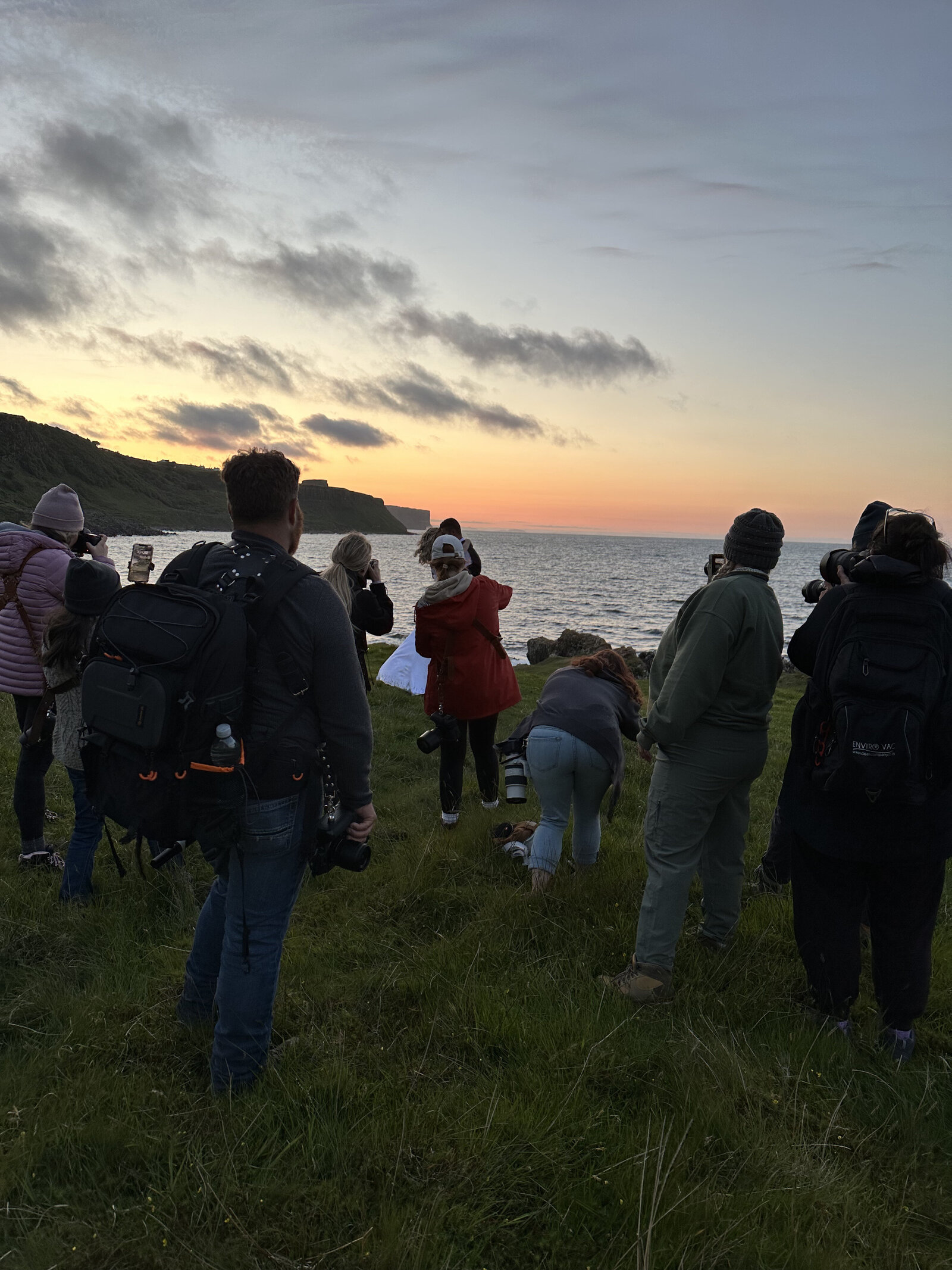 A group of people stand on grassy cliffs, watching the sunset over the ocean under a partly cloudy sky—capturing moments like at a California photography retreat for storytellers and soul seekers.
