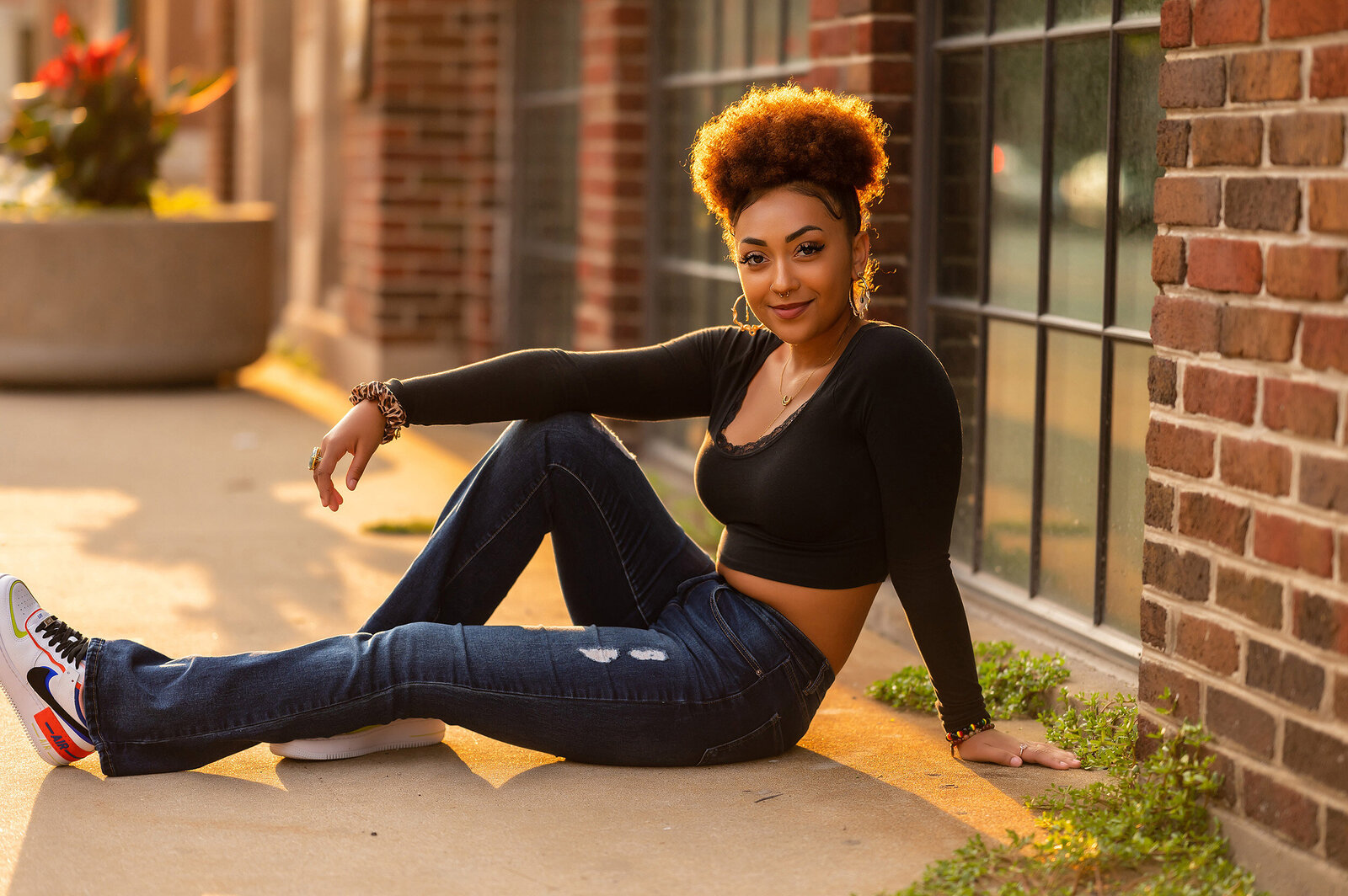 Backlit portrait of a New Berlin area high school senior seated on a sidewalk in Milwaukee's Third Ward. She is Black with brown curly hair and wearing jeans and a black long sleeve crop top.