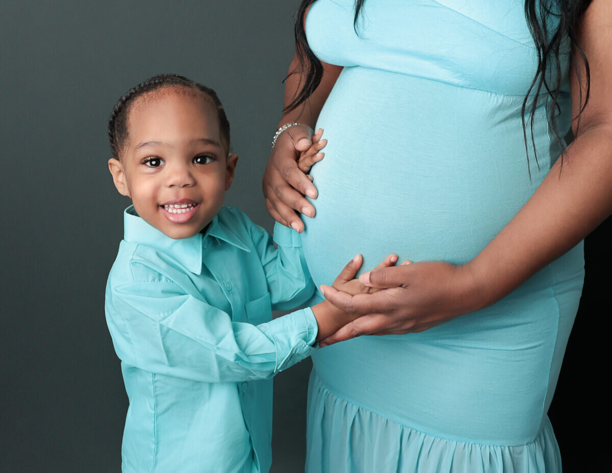 A son gently holds his mother's pregnant belly during a studio maternity photography session in Rochester, NY. The warm lighting highlights the joyful bond between them, capturing the essence of family love and anticipation.
