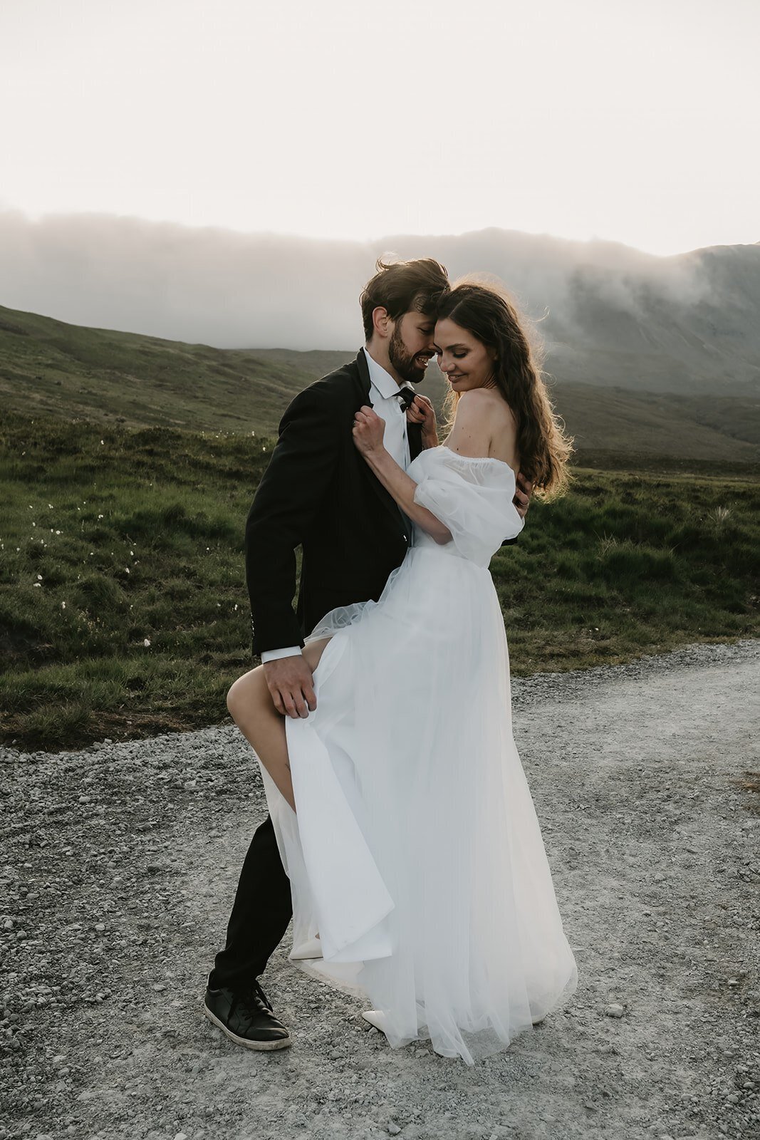 A bride and groom embrace on a mountain path at sunset, with the bride lifting her dress slightly—a moment evoking the wild-hearted spirit of an Oregon coast photography retreat for artists.