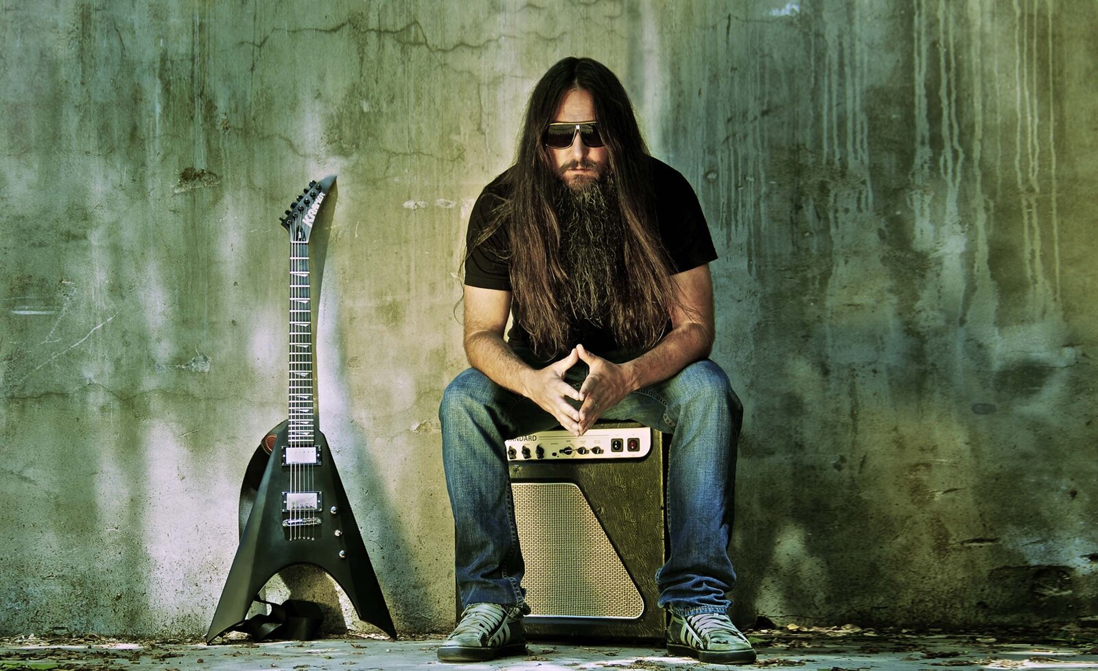 Musician portrait Rick Jackett sitting on amp beside black guitar cement wall backdrop