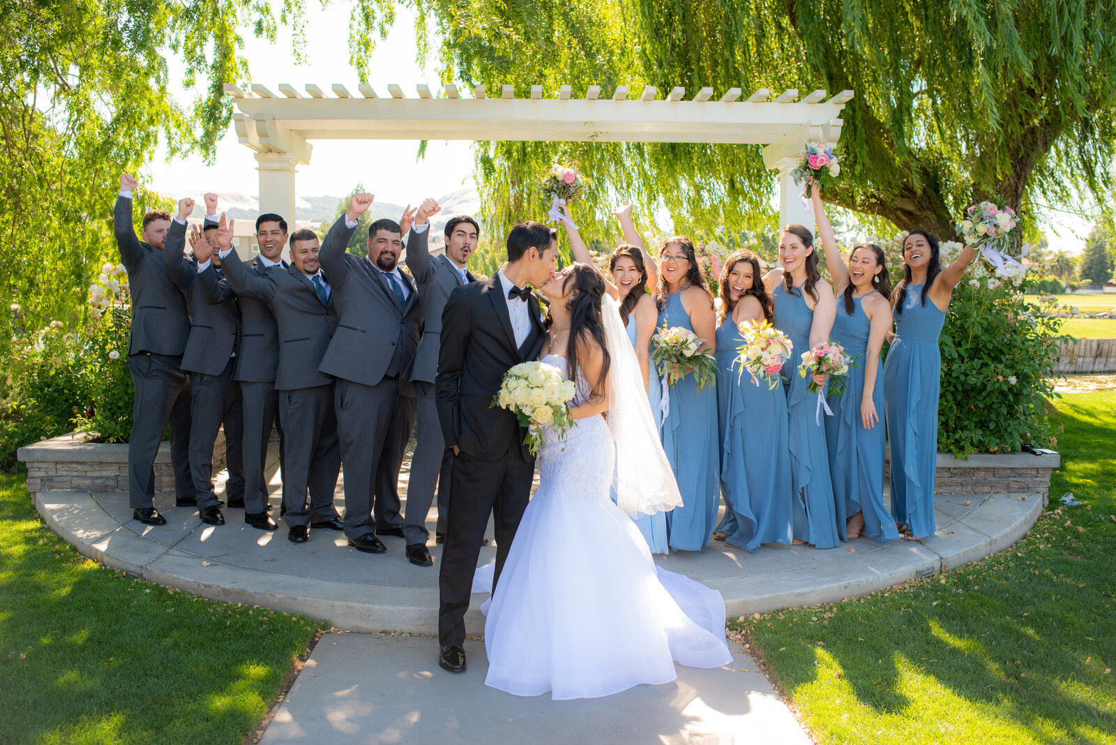 Bride and groom kiss in front of wedding party cheering them on from both sides. Girls are wearing a muted blue color and guys are wearing grey.  They are standing in front of a gazebo and large willow tree. Photo by wedding photographer philippe studio pro who has a studio in sacramento.