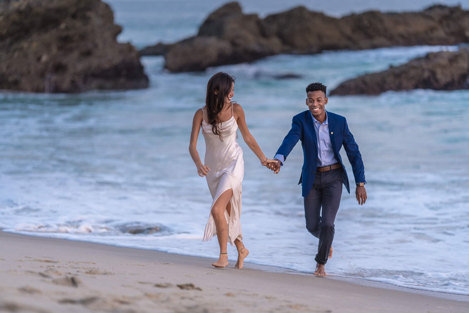 Young couple running from waves on beach while wearing formal clothes.