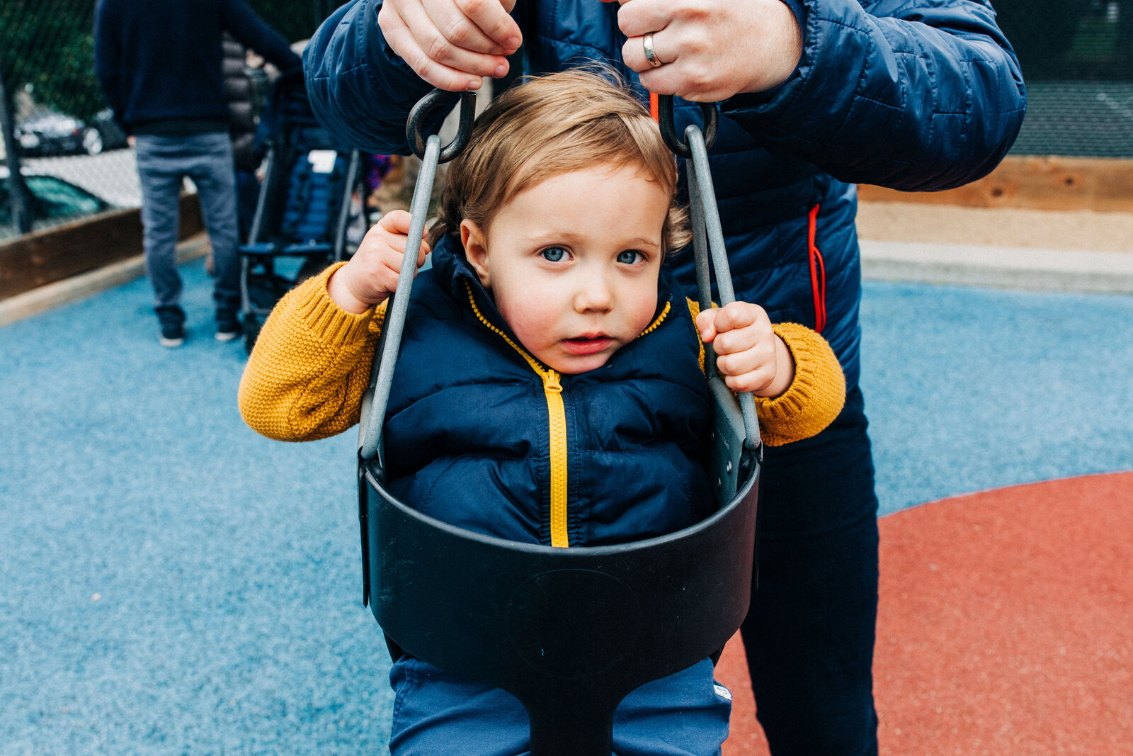 toddler boy in yellow sweater and navy puffy vest sitting in a baby swing at the park