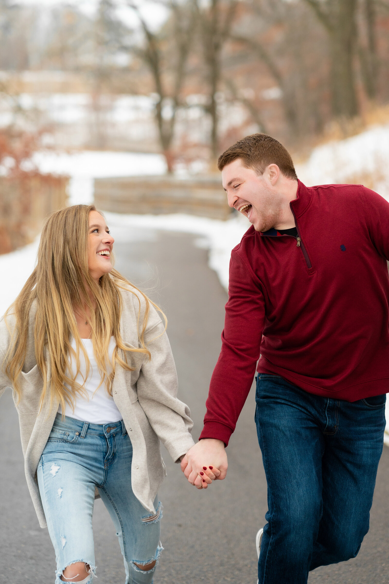 Man and woman in sweaters and jeans laugh while holding hands in the snow.