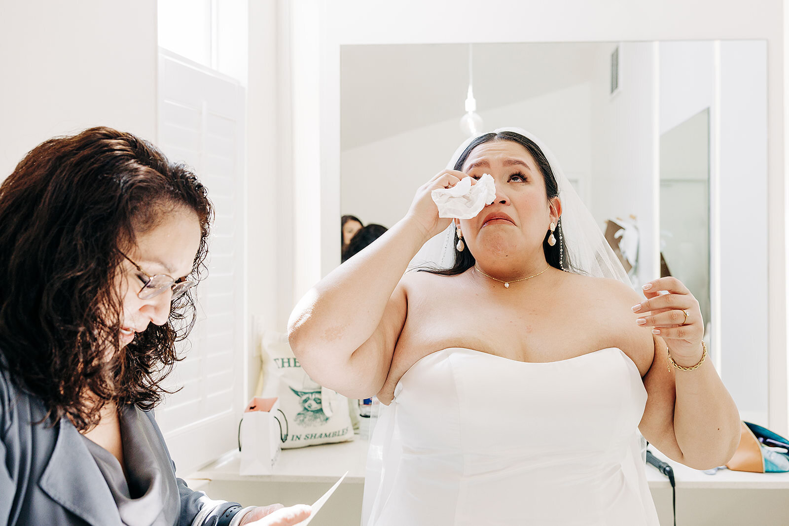 A bride in a white dress wipes tears from her eyes with a tissue, while a woman beside her reads from a piece of paper in a bright, softly lit room, beautifully captured by a Denver Wedding Photographer.