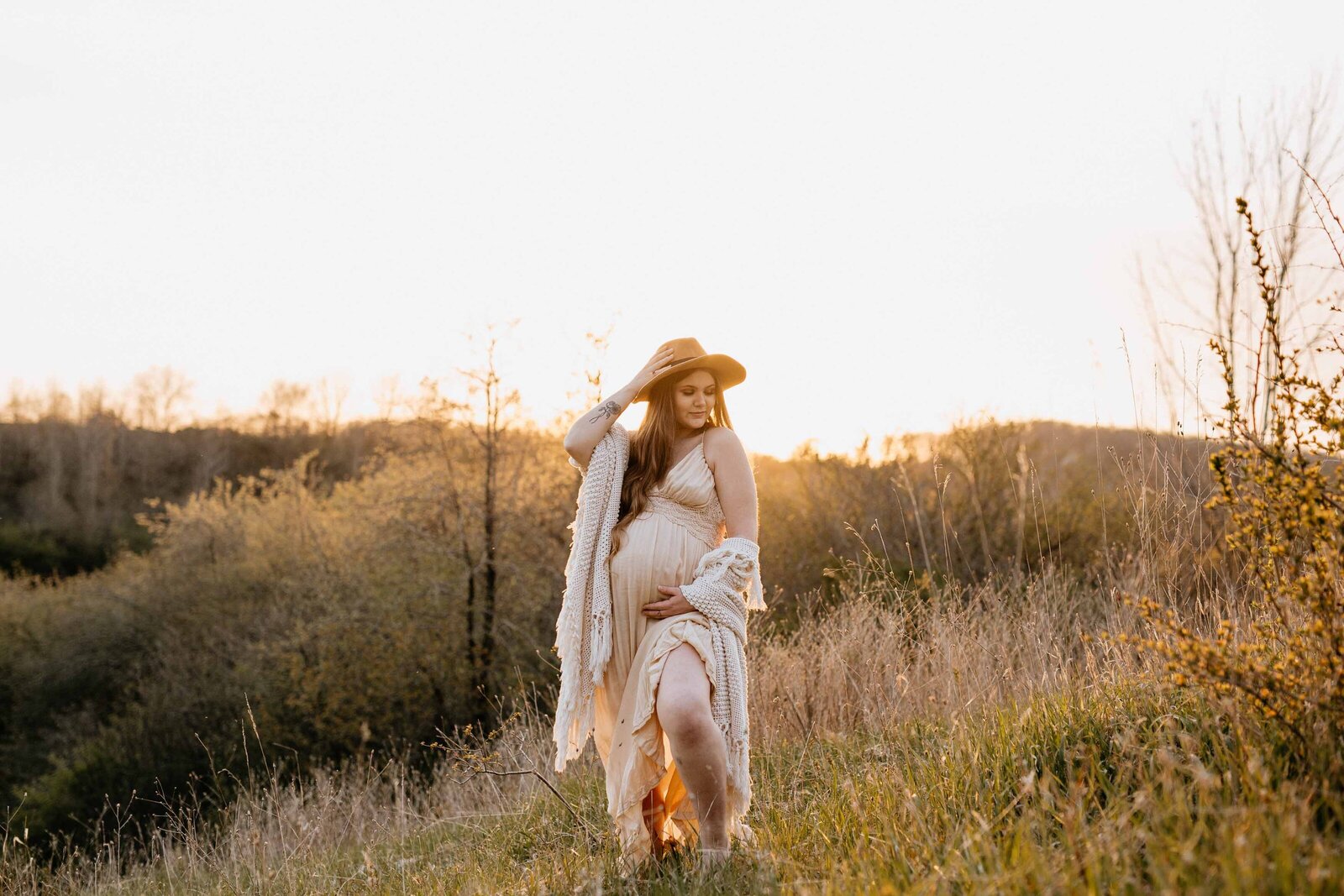 Expectant mom standing for portraits in London, Ontario field with rolling hills in the background. Mom is wearing a long cream dress with her left and under her bump and her right hand adjusting a wide-brimmed felt hat. Shot at golden hour with warm light.