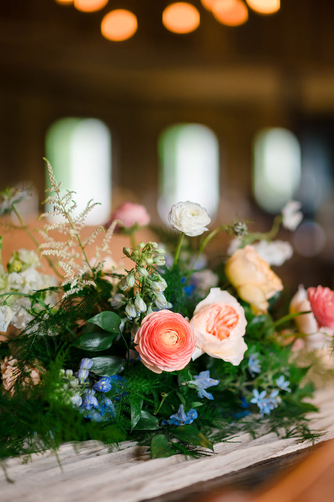 A close-up of a rustic chic floral arrangement on a wooden table, featuring pastel roses, white blossoms, and blue accents