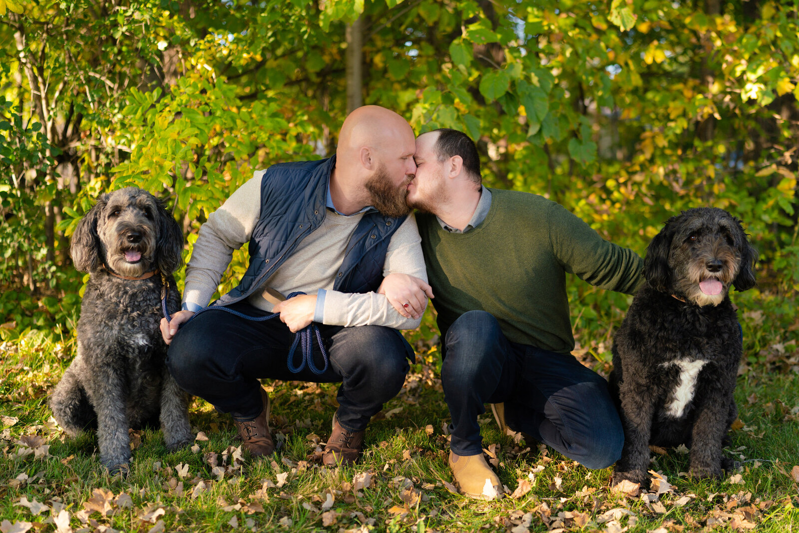 Two men kiss with their dogs sitting on either side of them.