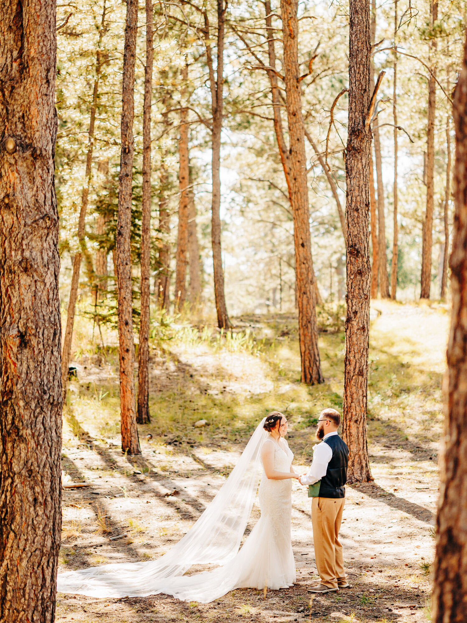 A couple stands in a forest in Colorado wearing their wedding attire. The bride has a long veil and a long train on her dress. The groom wears a green vest.