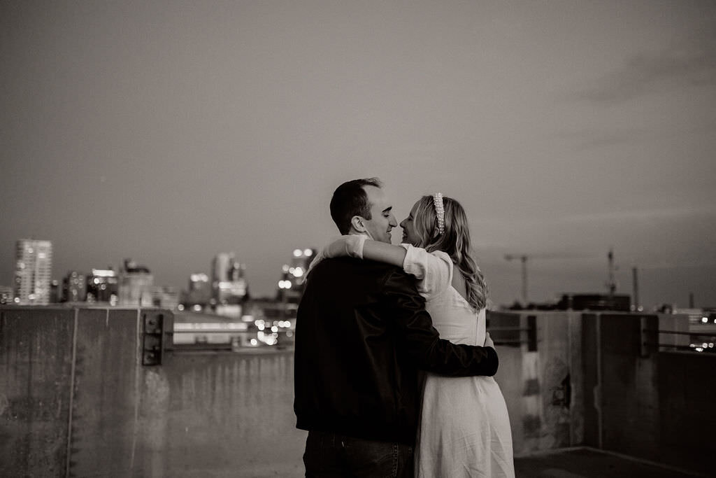 Gorgeous vintage black and white engagement photos by Morgan Ashley Lynn Photography on a rooftop in Milwaukee with man and woman with arms around eachother, looking at eachother smiling, woman has a pearl headband on, vintage dress and vintage gloves up to her elbow