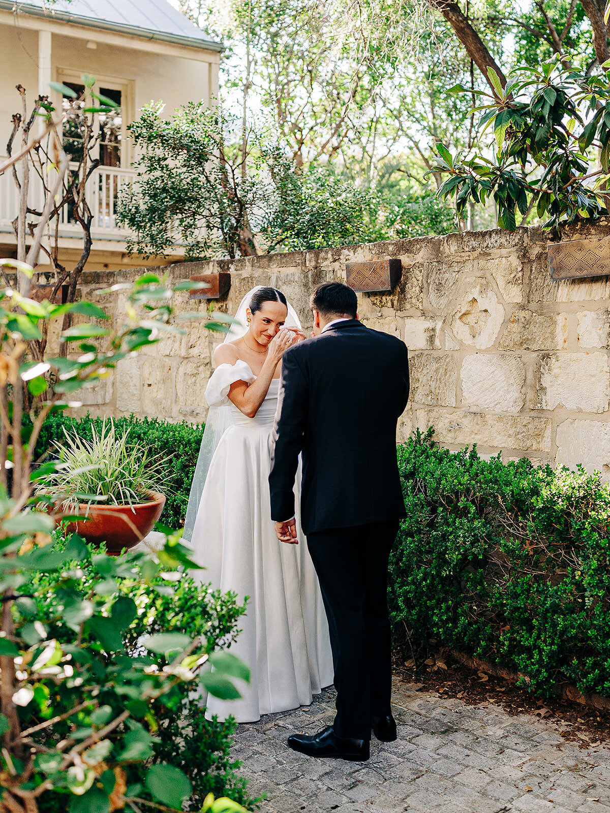A bride in a white dress wipes her eyes while standing with a groom in a black suit in a sunlit garden, surrounded by greenery and a stone wall, beautifully captured by a Denver Wedding Photographer.