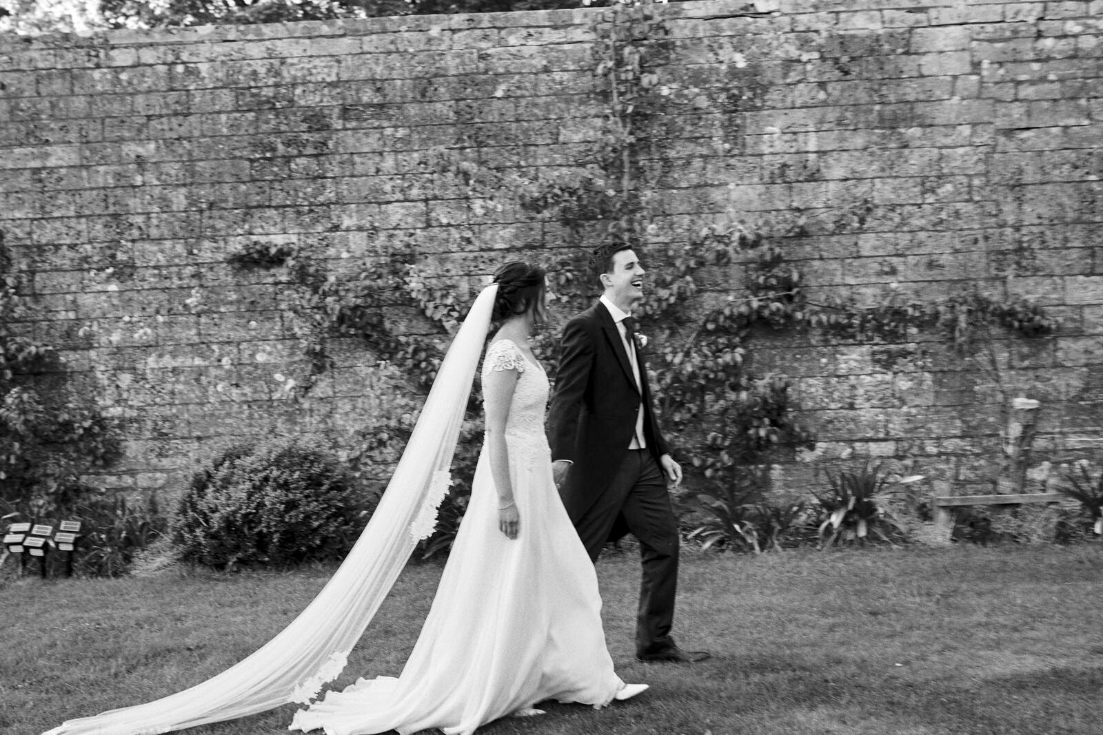 Black and white photo of bride and groom walking past stone walled garden