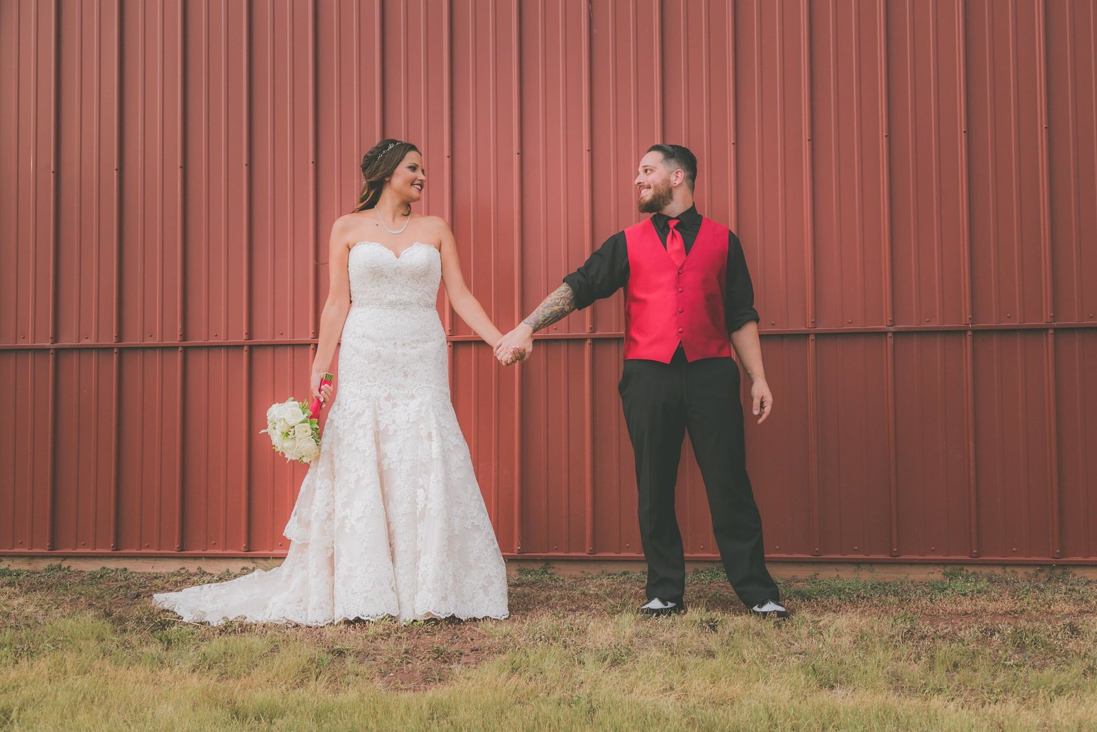 Bride and groom hold hands and look at one another against red barn background.
