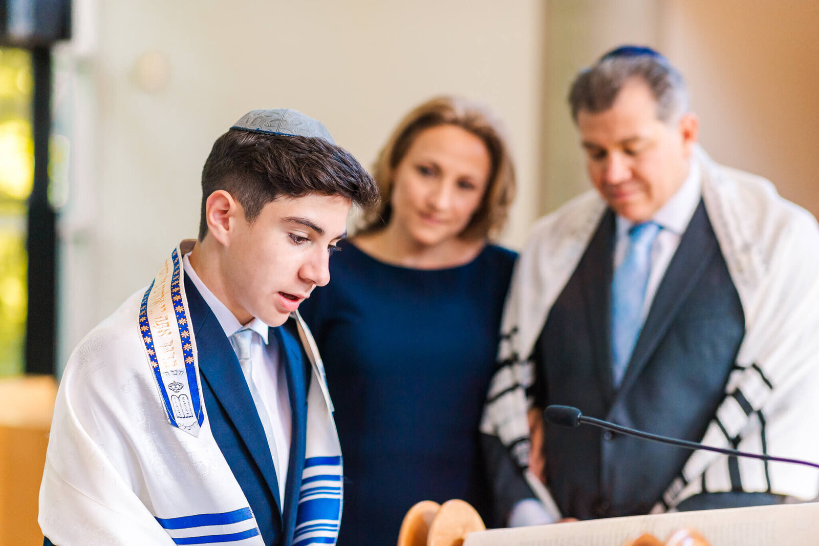 A teen boy in a blue suit and tallit performs the Haftarah with mom and dad looking on in an image in Bellevue Bar and Bat Mitzvah Photography