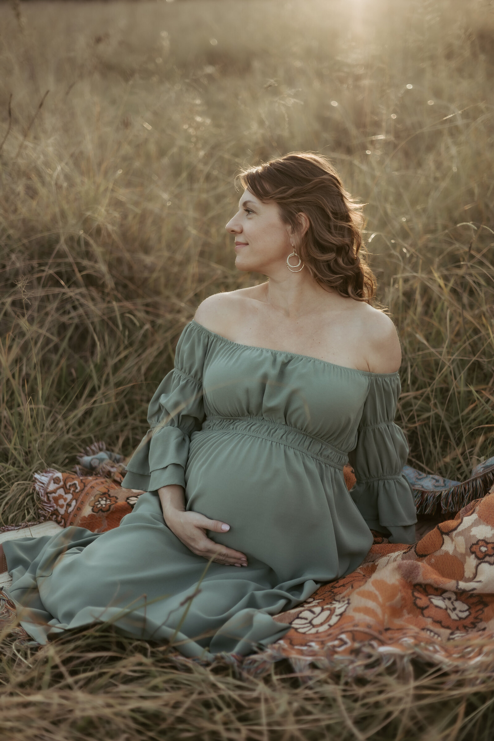 Stunning pregnant woman in off the shoulder green dress sitting in a lovely field holding her bump while the sun glows in her hair like a halo