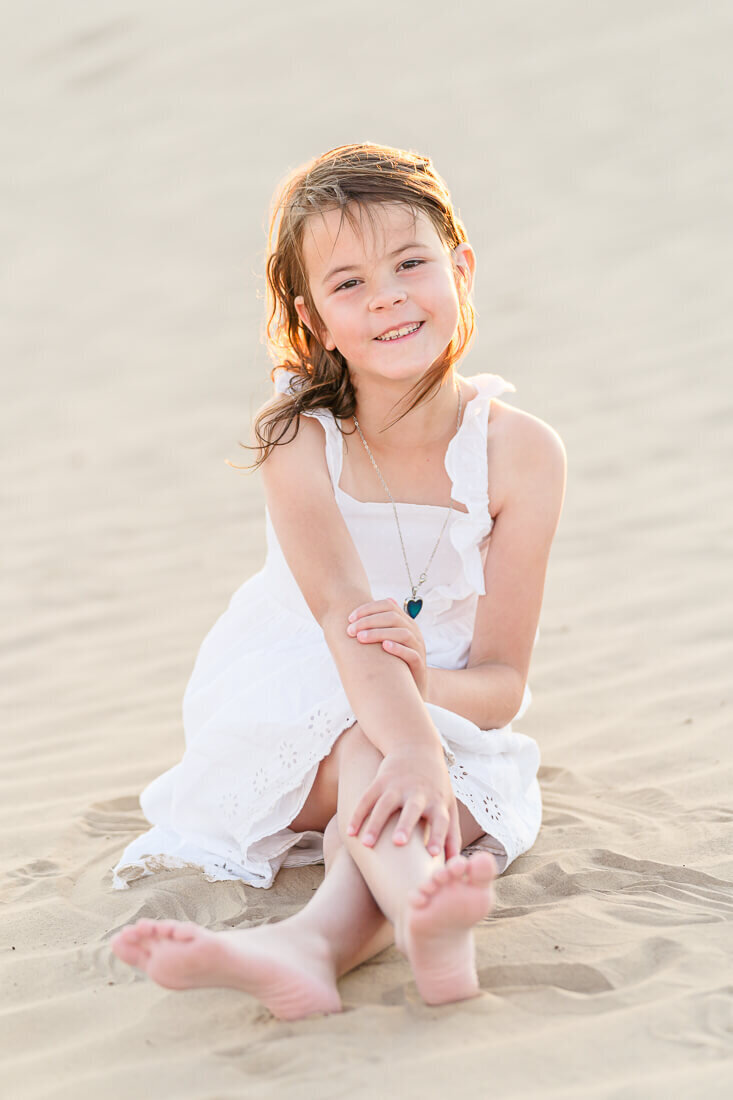 8 year old girl wearing a white eyelet lace dress while sits in the sand at Little Sahara Recreation Area in Nephi. Captured by Salt Lake Photographer Melissa Woodruff Photography.