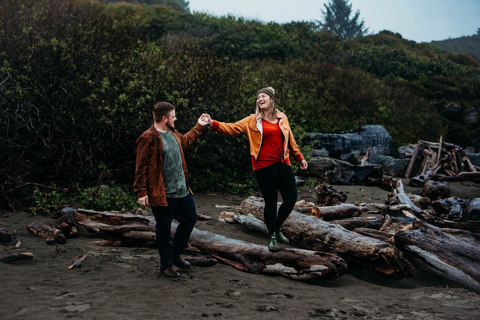 Couple strolls on beach while walking over fallen logs