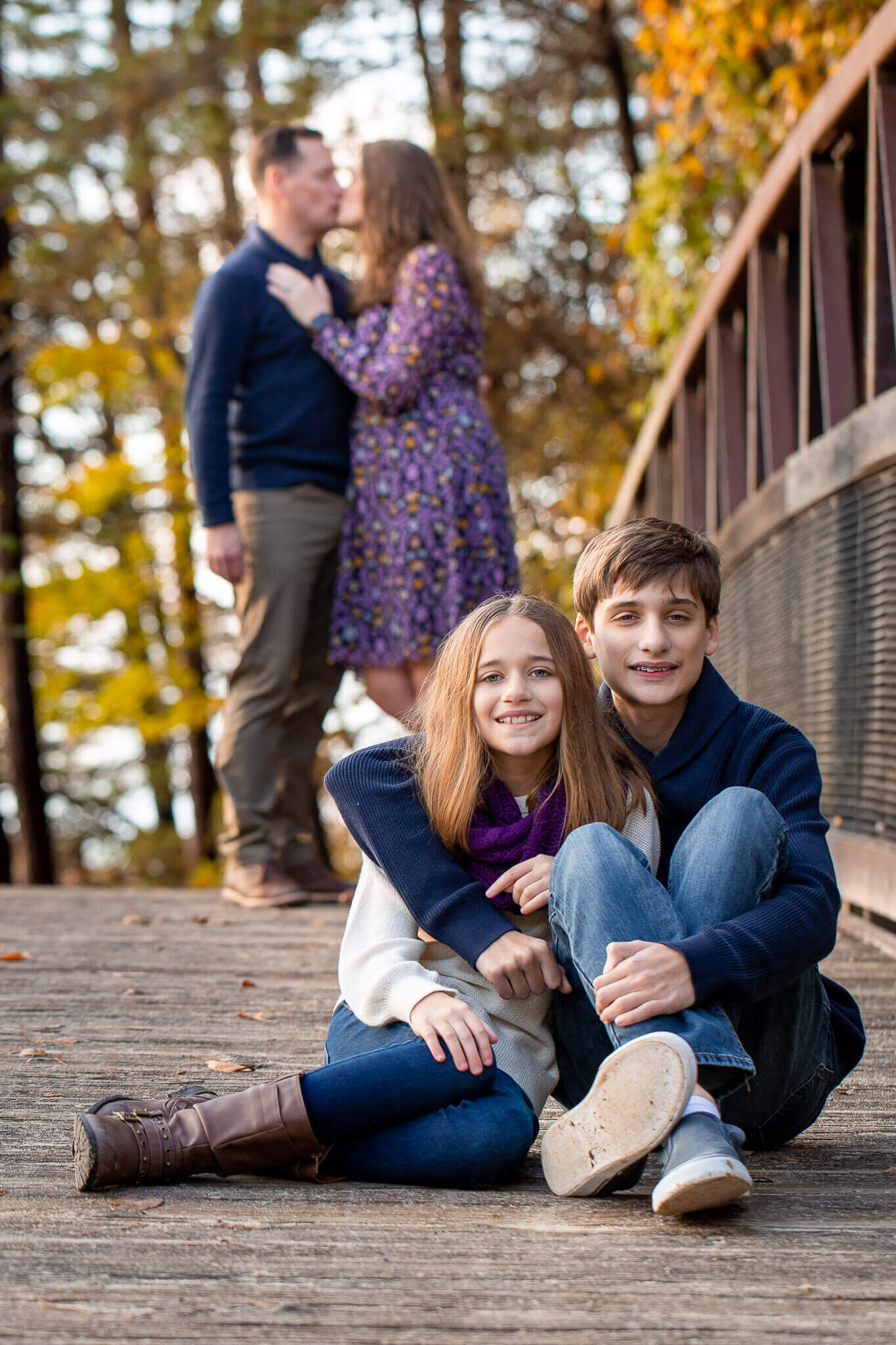 Siblings sitting on a bridge while their parents stand behind them kissing during their Springfield family photography session.