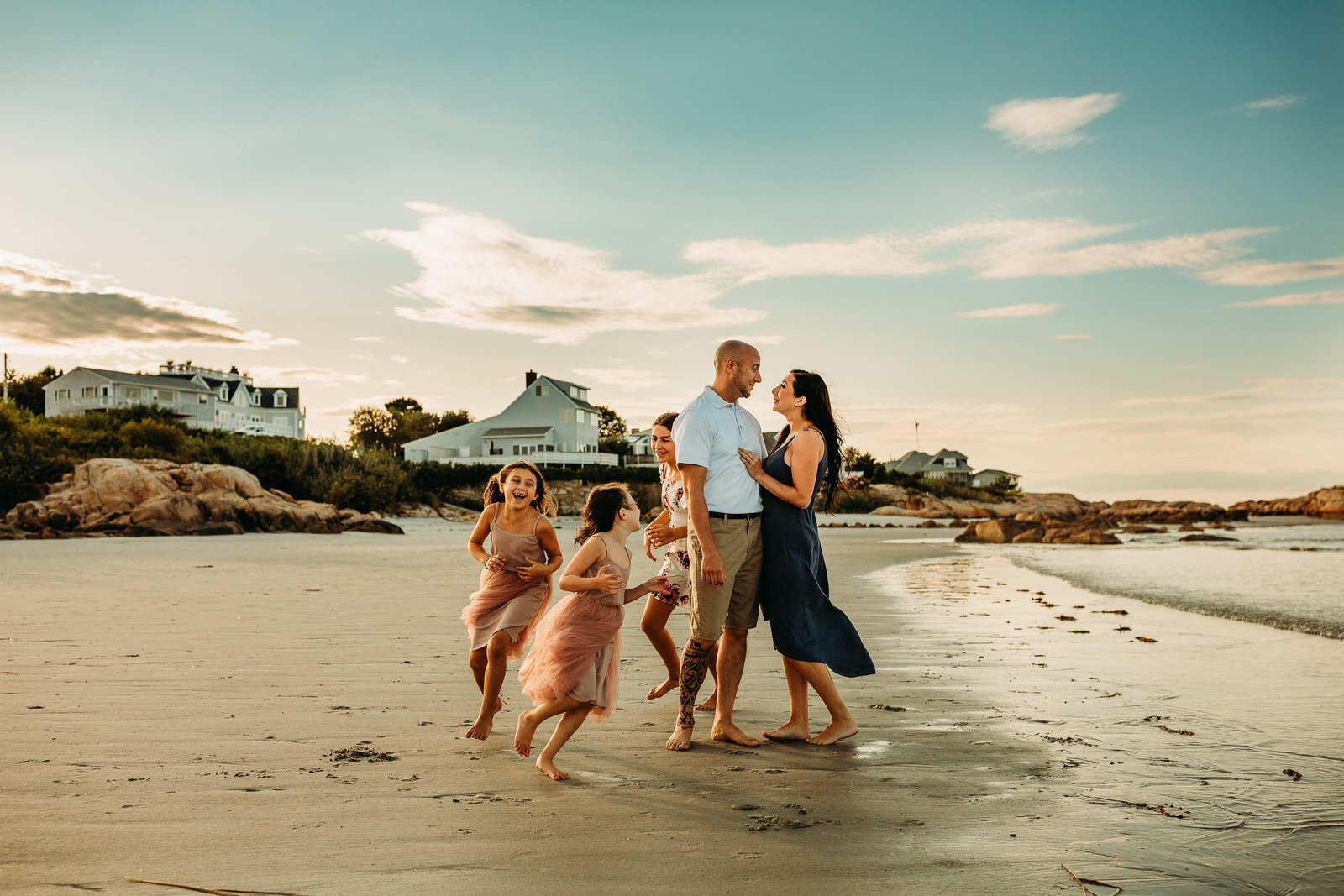 couple stands together on beach while daughters run around in family photo session