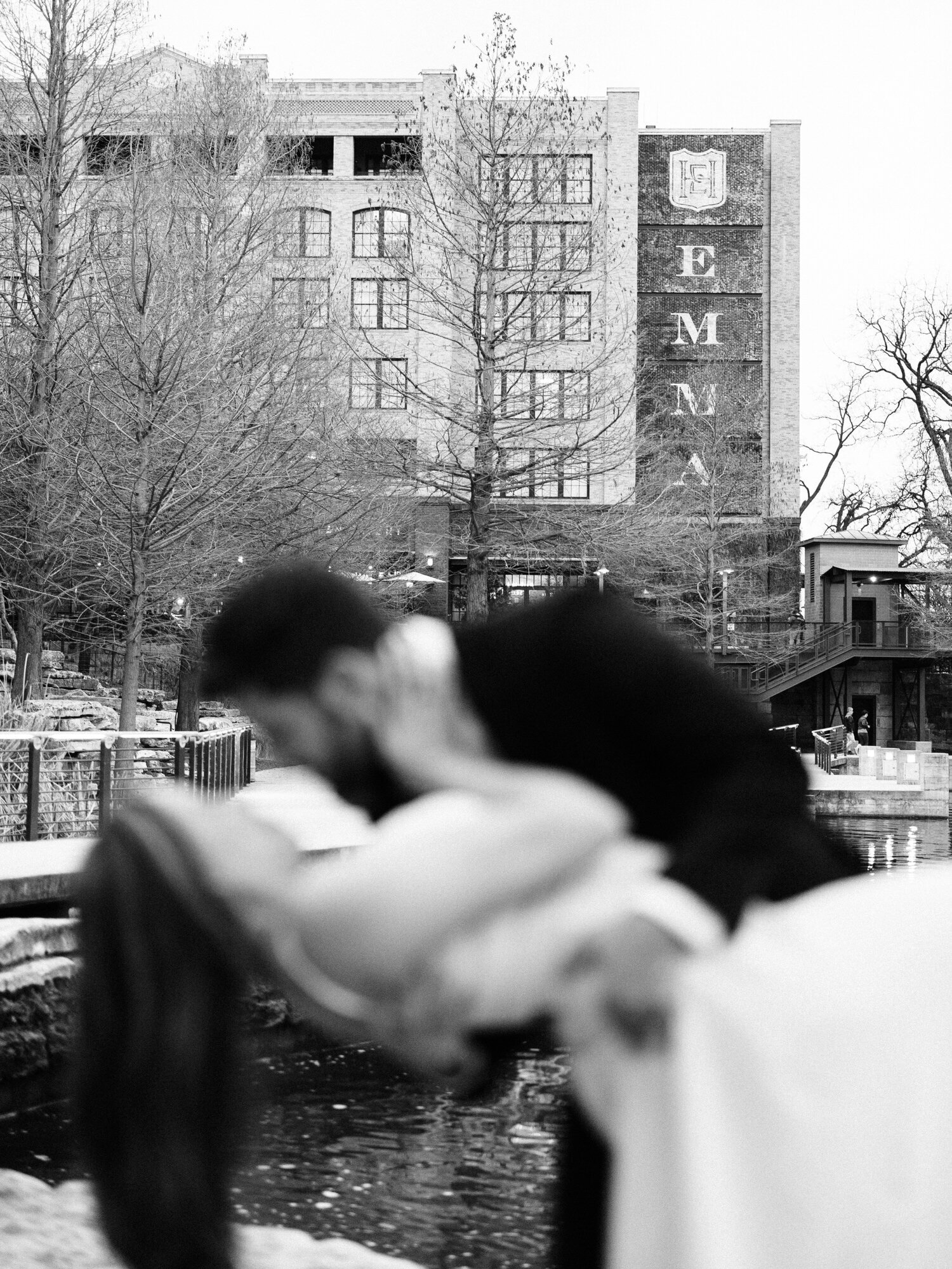 This image, taken at the Pearl in San Antonio, TX, features Hotel Emma prominently in the background, with a couple kissing in the foreground. The couple is out of focus and the man is dipping the woman.