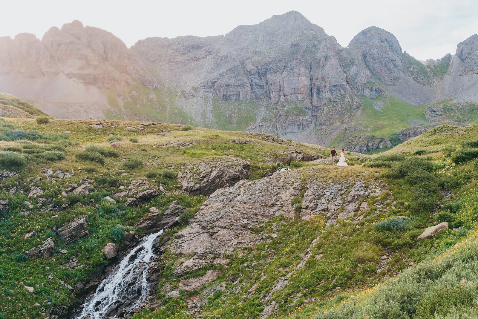 Wedding couples walks above a waterfall streaming down a grassy mountainside.