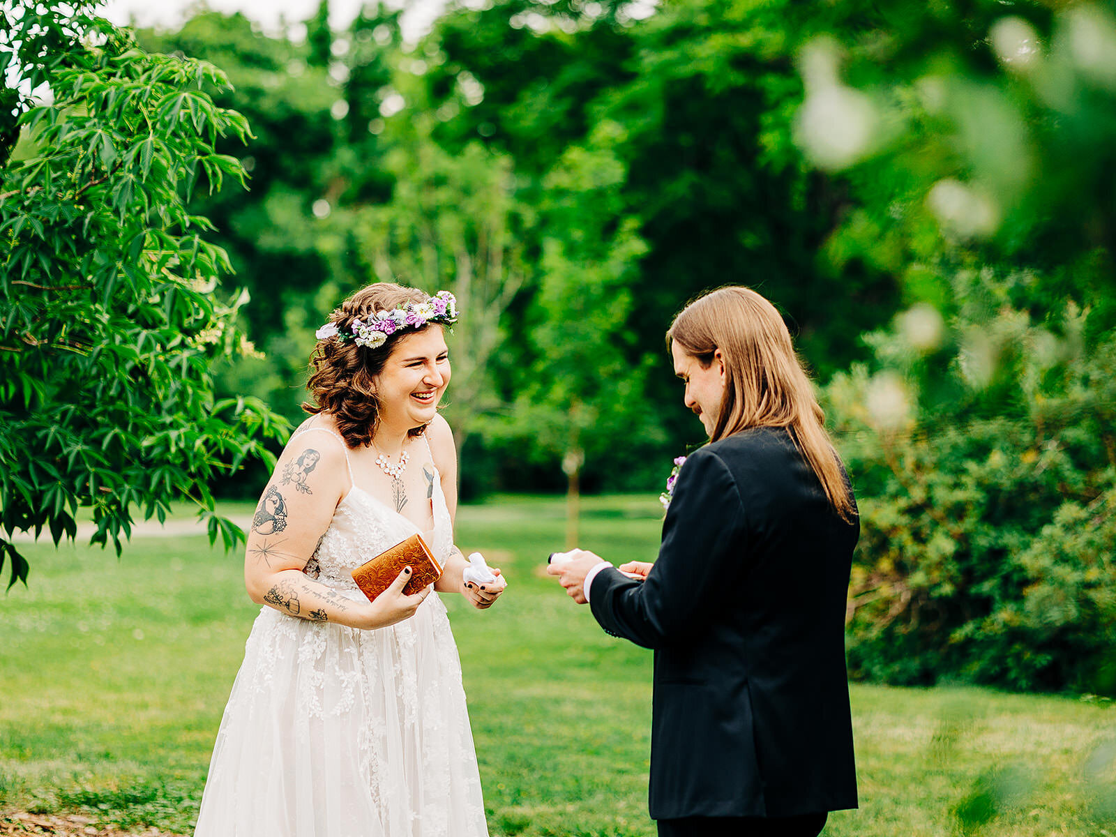 A bride in a white dress and floral crown smiles while holding a small clutch, facing a person in a black suit with long hair, as they exchange vows outdoors in a lush park—captured by your Colorado Wedding Photographer.
