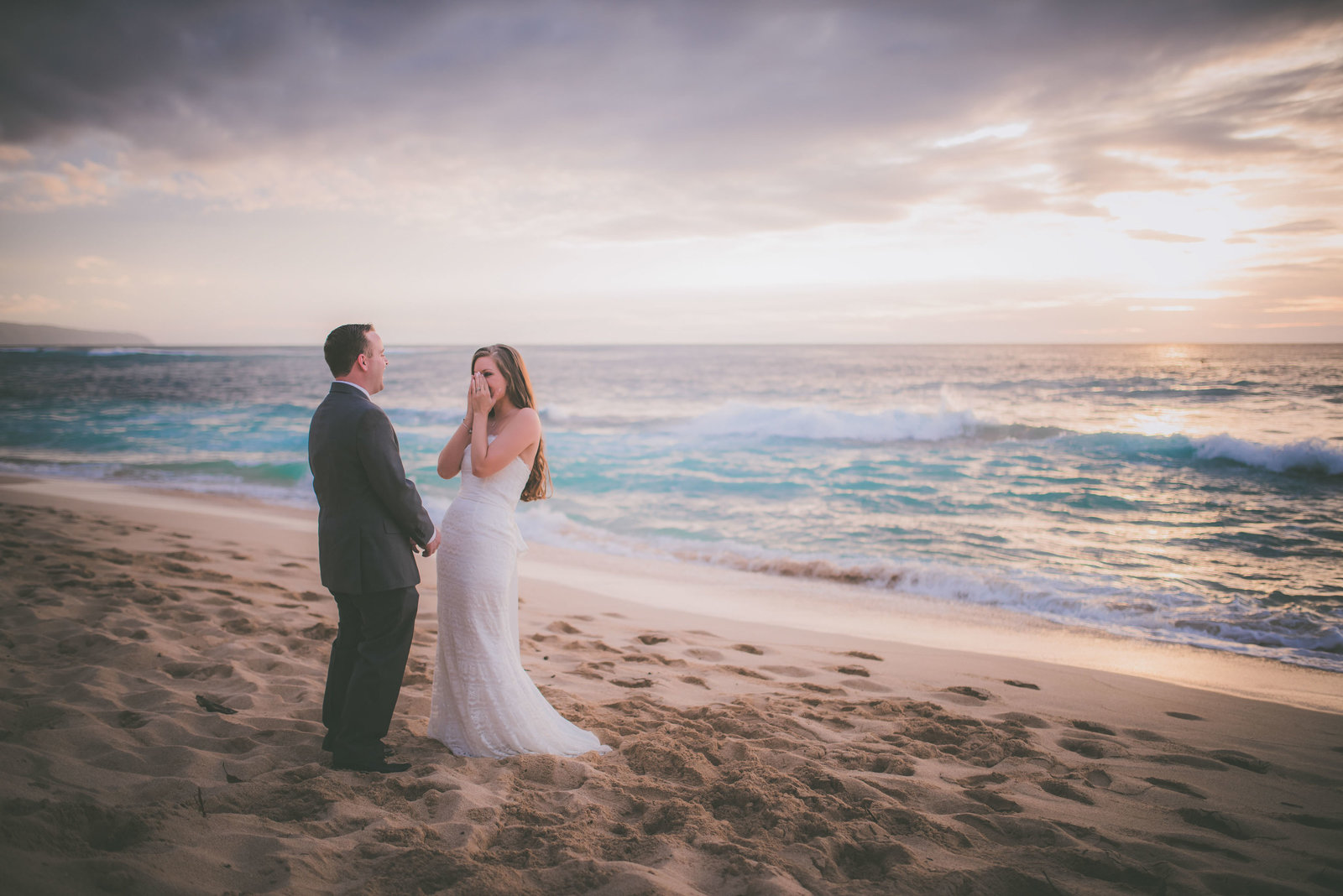 Bride places hands on her face while looking at groom on Oahu beach.