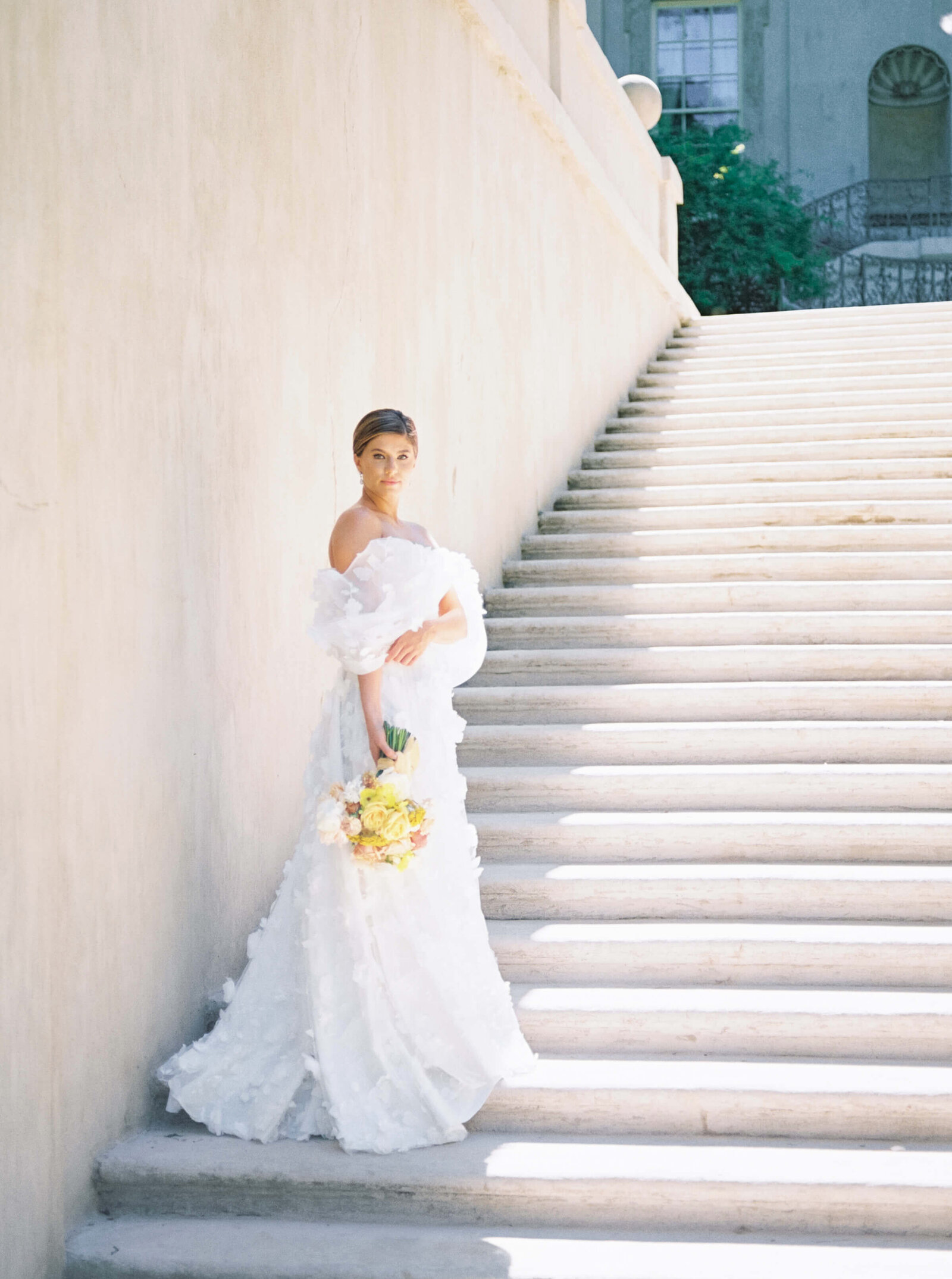 Bride stands on white stone stairs in designer wedding dress while holding yellow bouquet