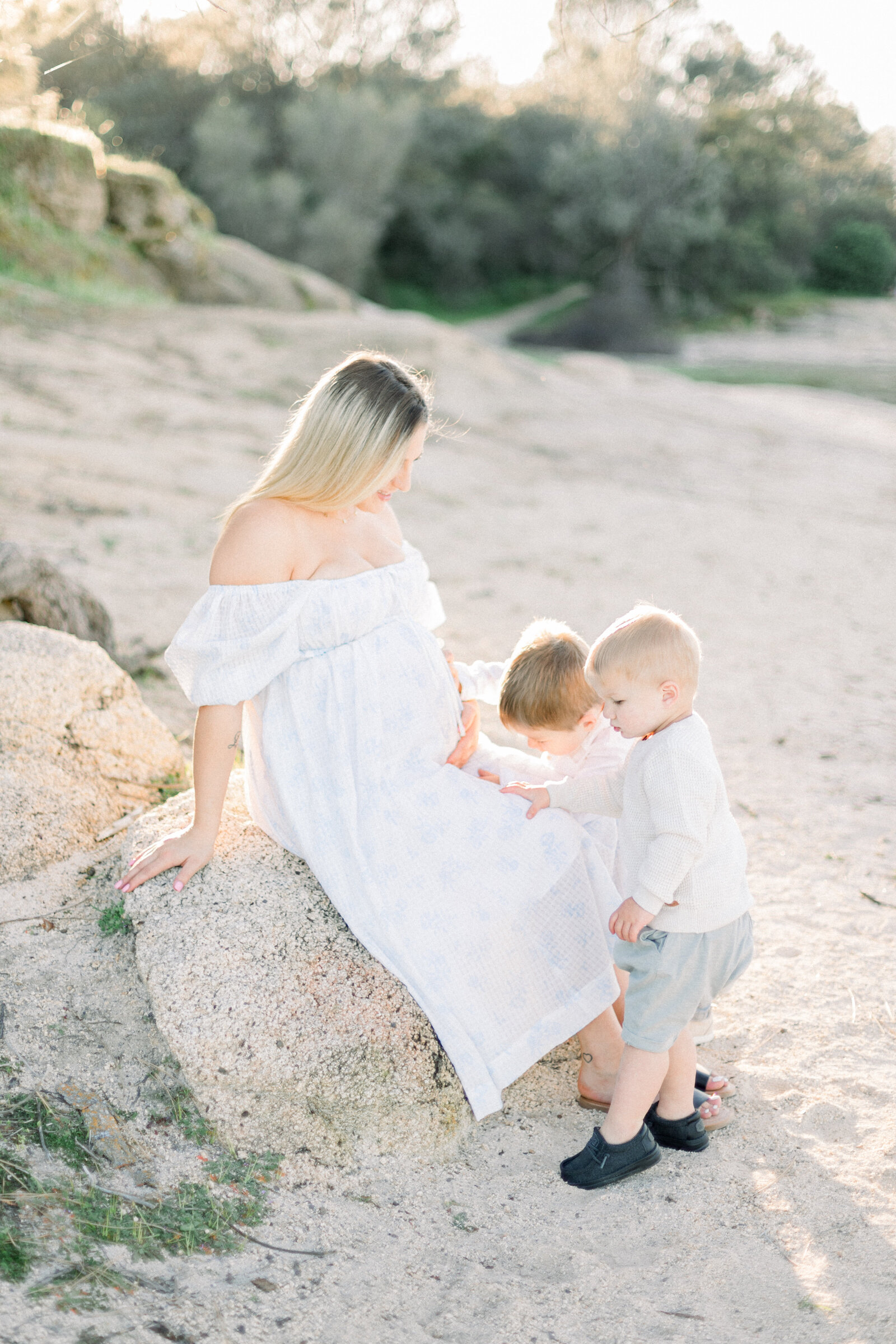 Image of expecting mother in white dress sitting on a rock with her two sons looking at belly taken by sacramento maternity photographer Kelsey Krall