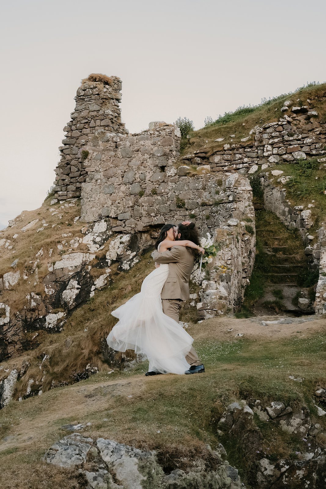 A couple in wedding attire kisses in front of ancient stone ruins on a grassy hillside, capturing the romance perfect for an Italy photography retreat in the countryside for inspired storytelling.