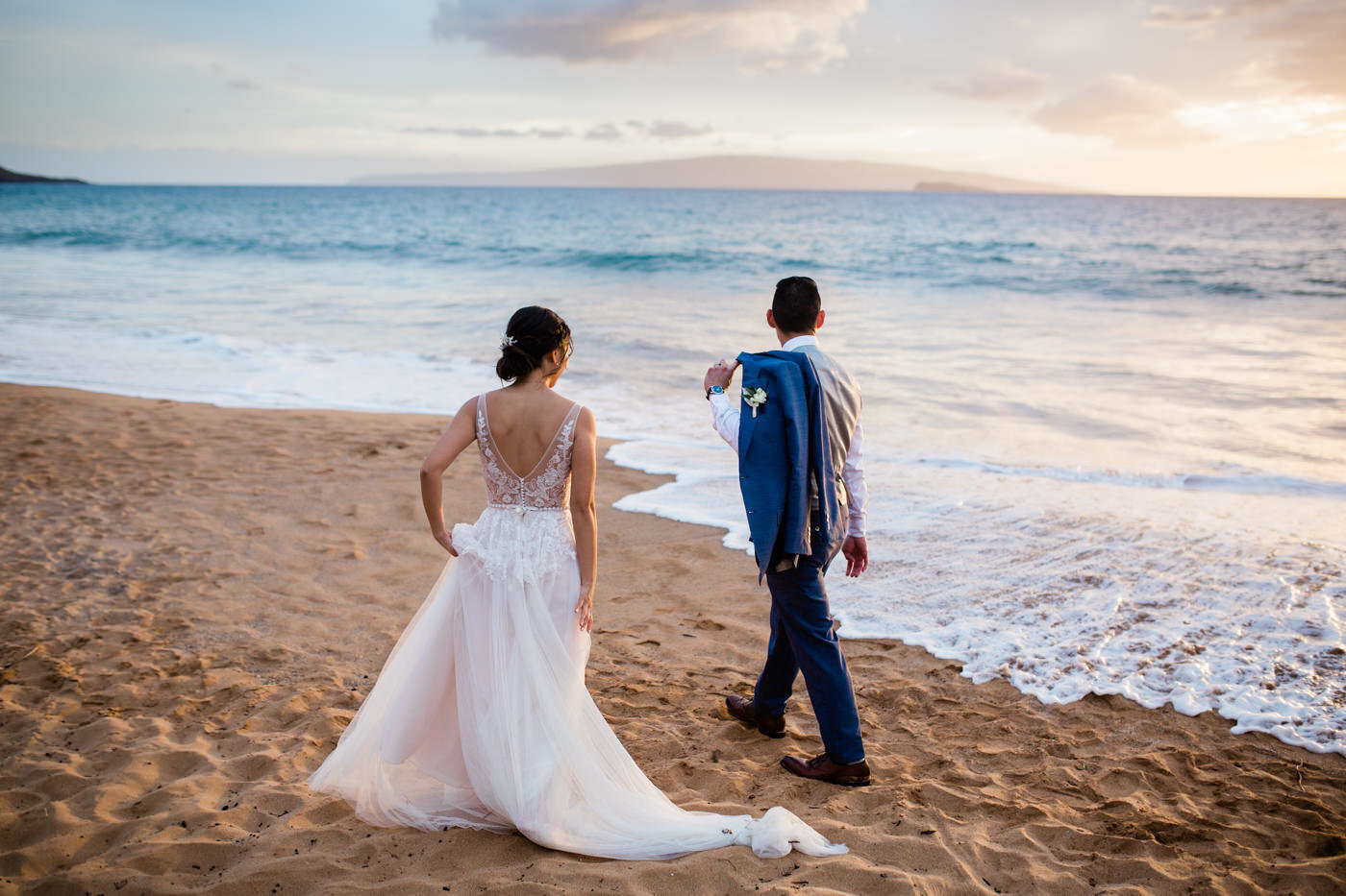 bride and groom on a beach in maui. The bride is wearing a gown by BHLDN and the groom has his suit jacket on the side of his shoulder. There is a pretty sunset.