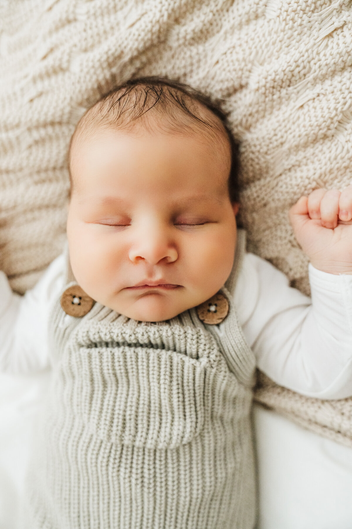baby in knit overalls lays on bed during newborn photos in boston