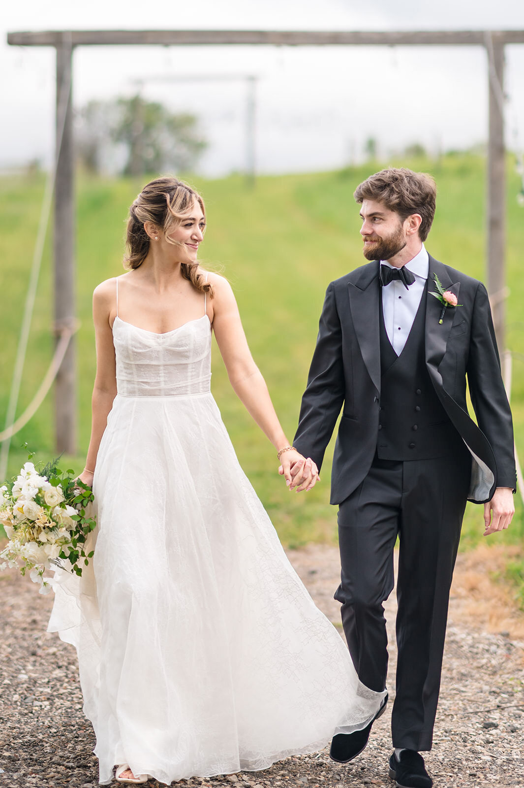 A bride in an elegant white gown and a groom in a sharp black tuxedo hold hands and smile at each other, walking under a rustic ski lift on a grassy field