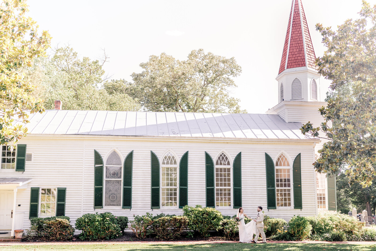 A bride and groom hold hands and walk together down a path beside the historic chapel where they had their Catholic wedding in Northern Virginia