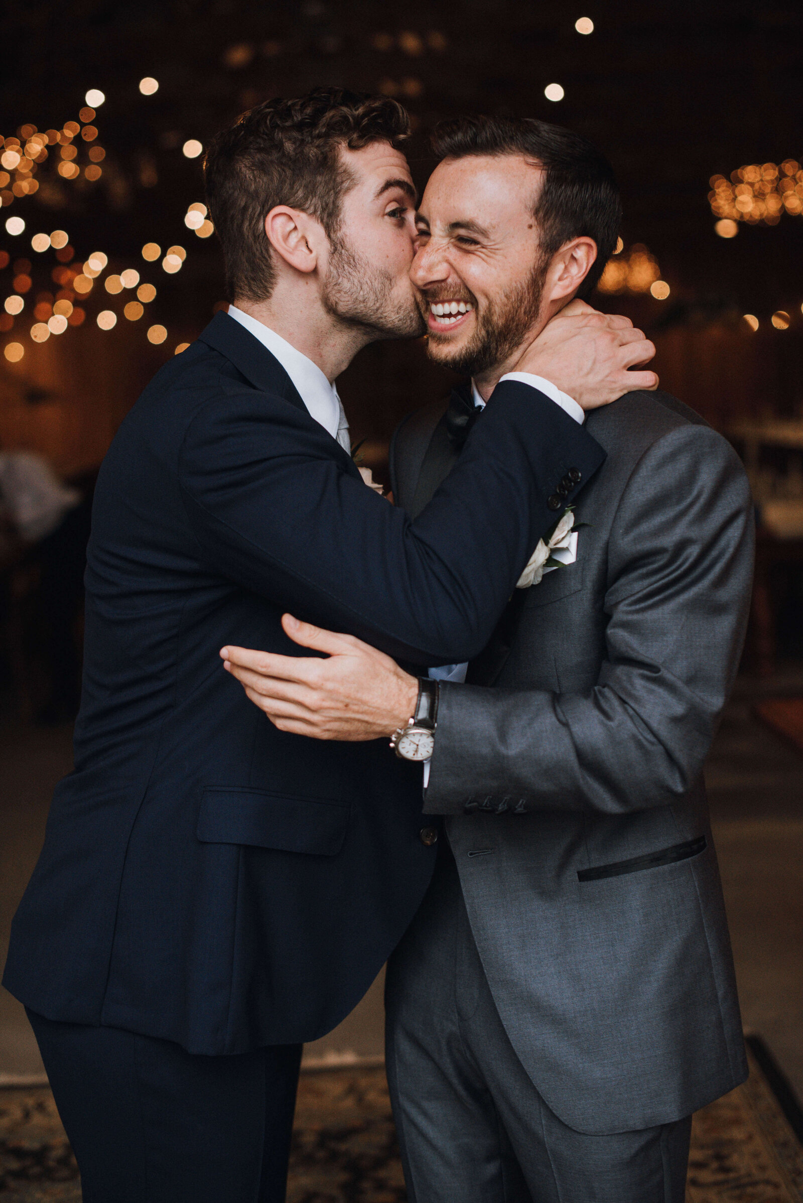 A laughing groom getting a playful kiss on the cheek from a groomsman