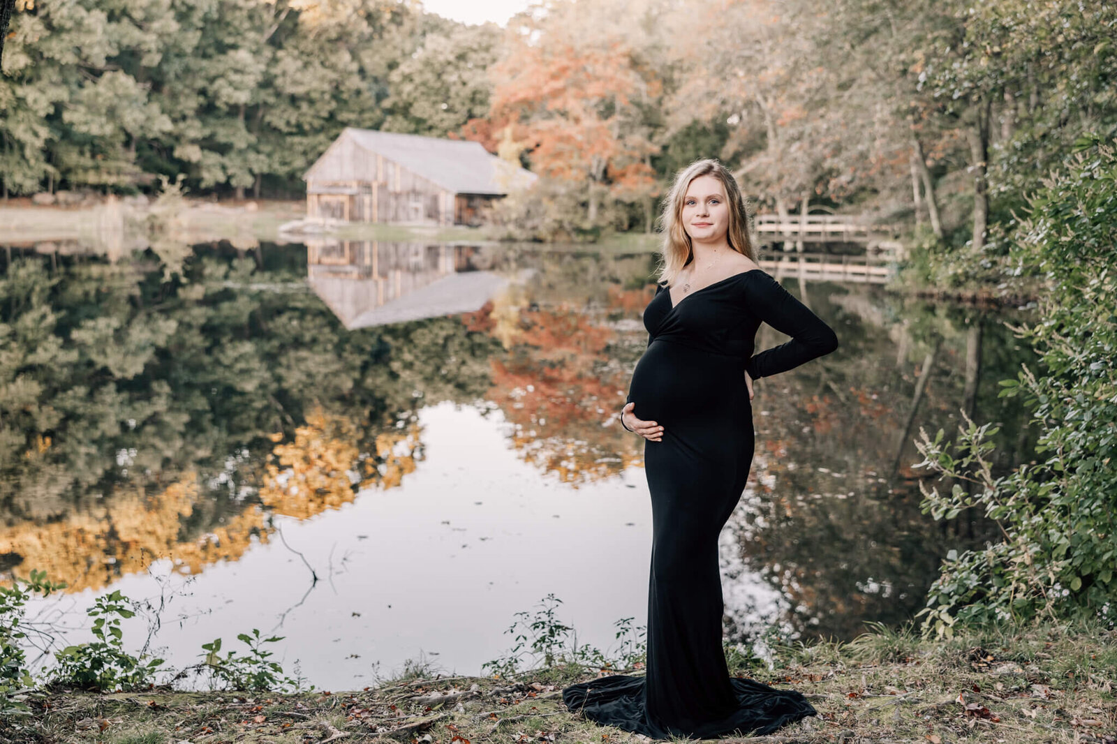 Pregnant woman with blond hair in black dress poses in front of Sawmill Pond in Ledyard, CT