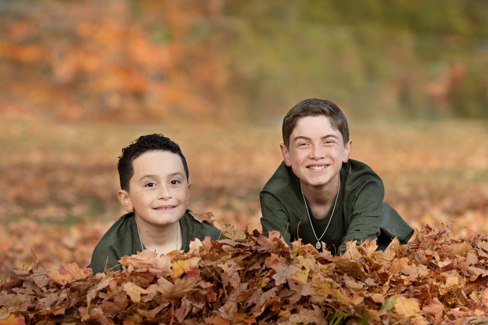 Two boys wearing green shirts laying in a pile of leaves smiling