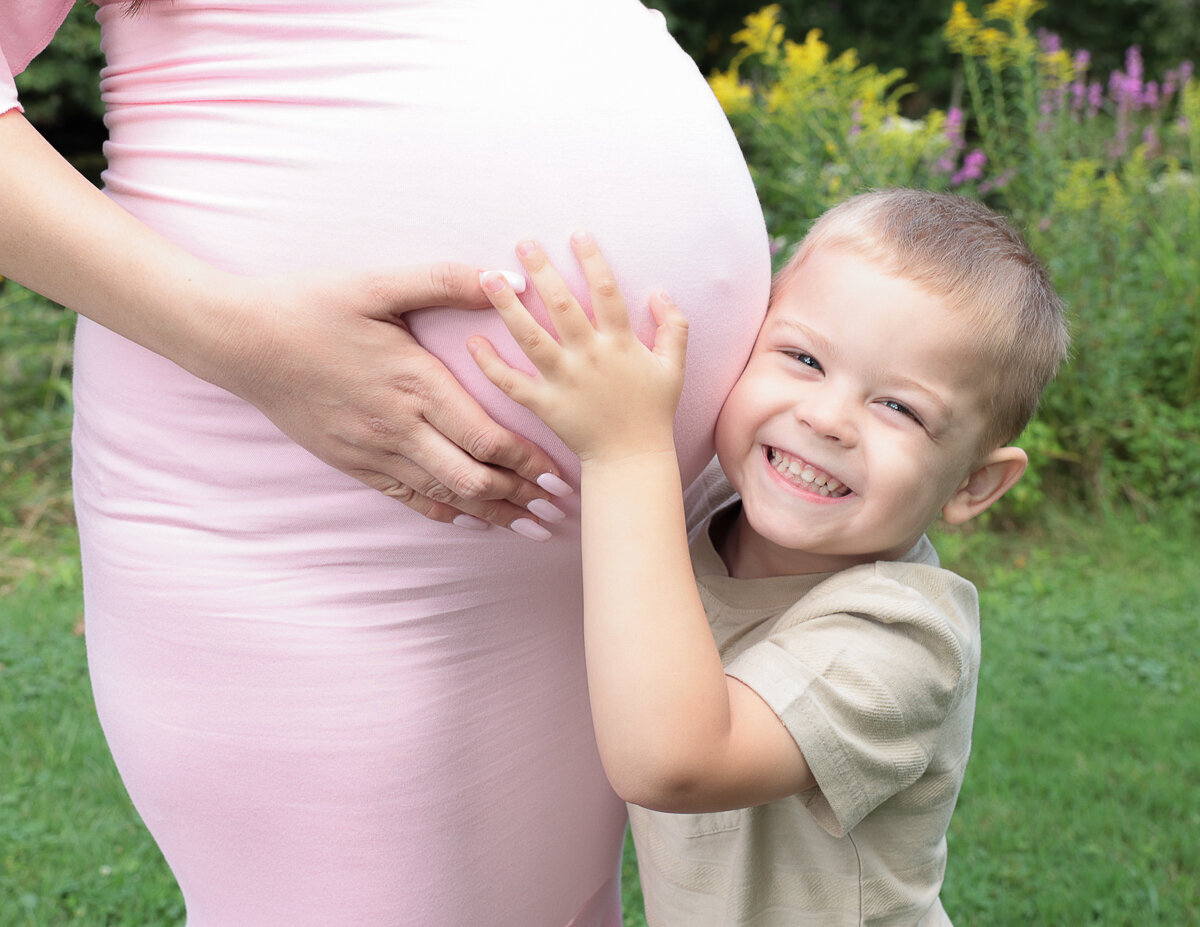A heartwarming outdoor maternity photograph capturing a young boy hugging his mother's baby belly, surrounded by the beautiful scenery of Rochester, NY.
