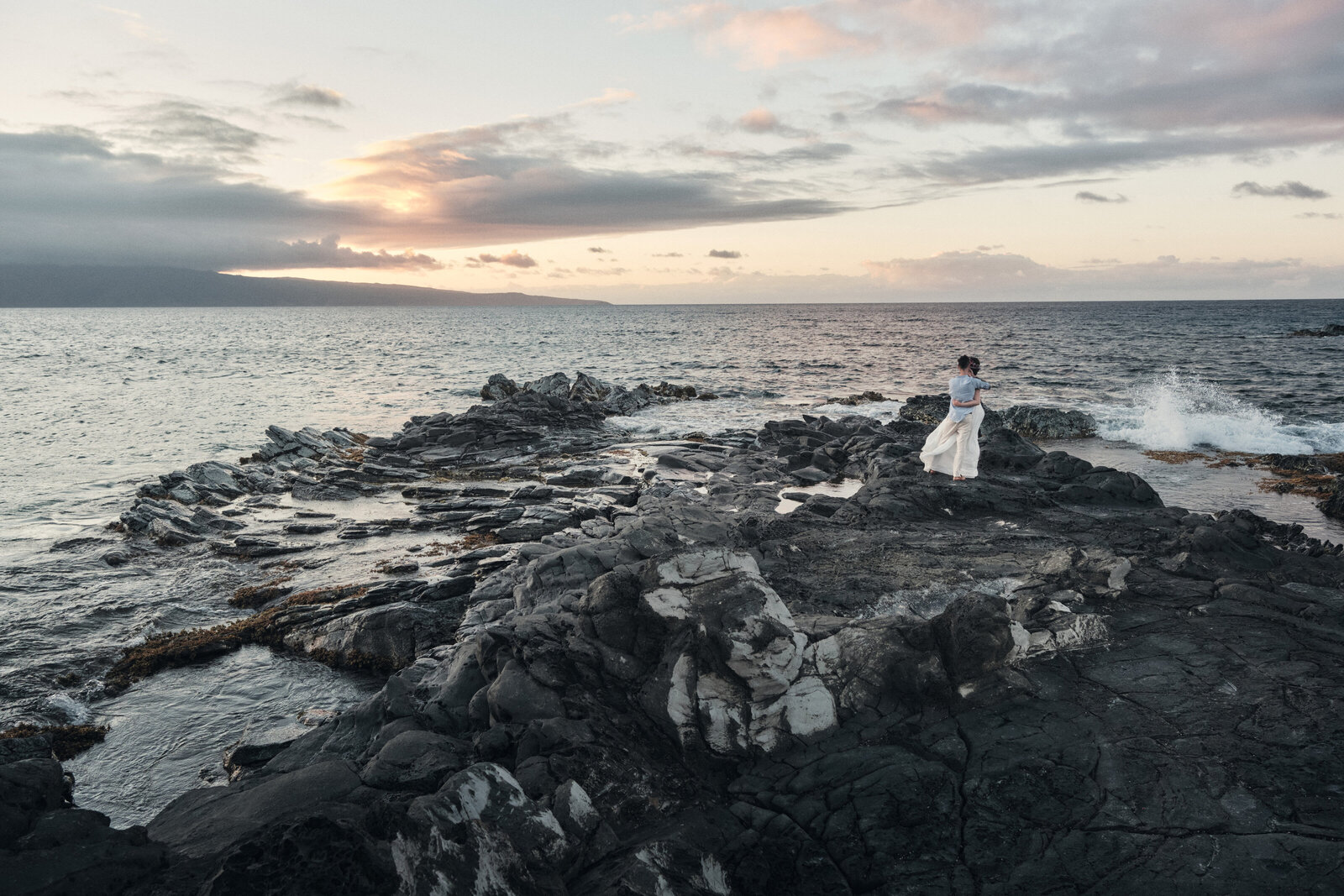 Water splashes upon rocky coastline as eloping couple looks on