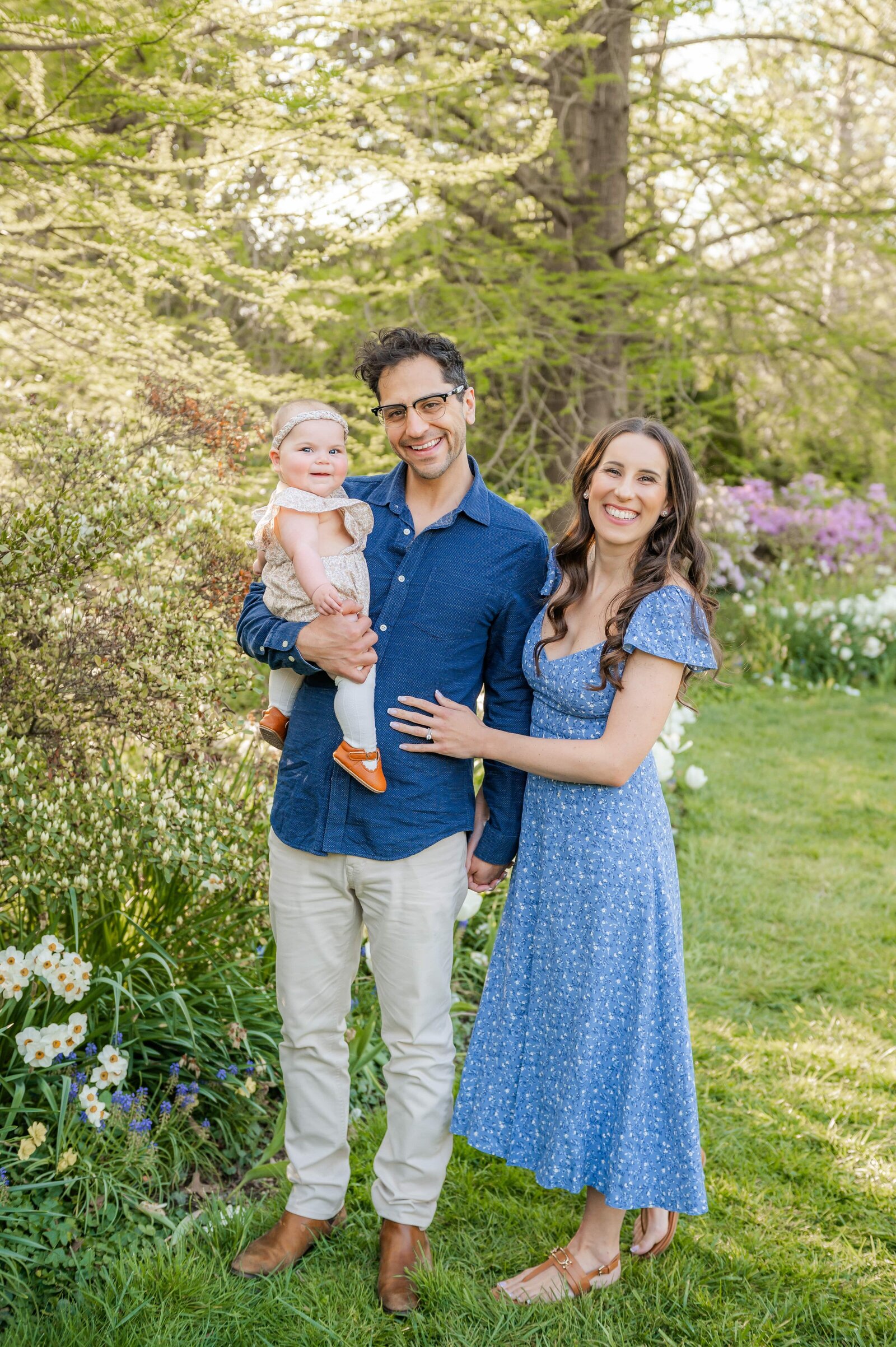 mom, dad and 6 month old girl smiling in a garden of flowers