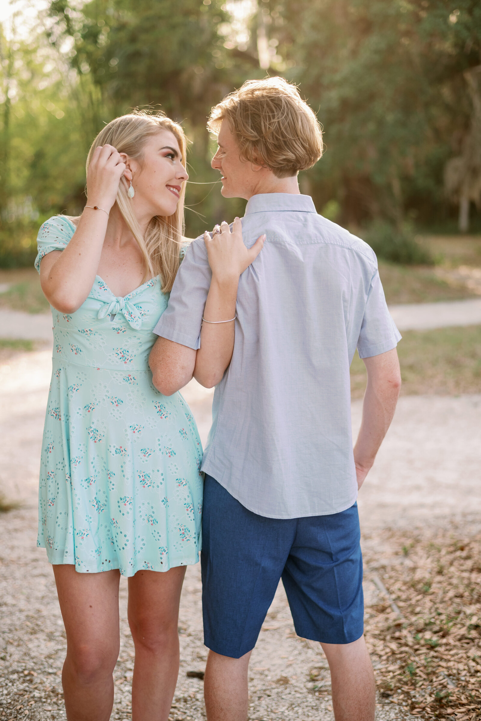 engaged couple standing opposite one another. She is facing the camera, looking at her fiancé while tucking her hair back behind her ear. He is standing with his back to the camera, looking at his faiancé. Their inner arms are folded together