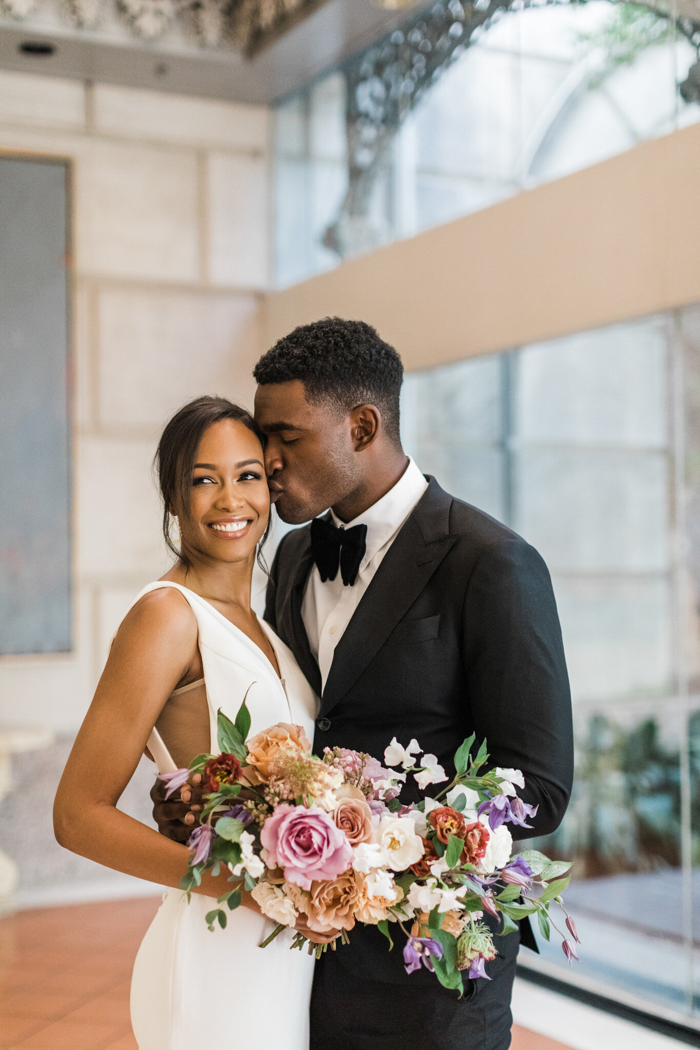 An African American bride and groom posing joyfully together at the Crescent Court hotel in Dallas, Texas. The bride is wearing a sleeveless white dress and holding a large bouquet while smiling and glancing of to the side. The groom is wearing a black tuxedo while kissing the bride on the cheek with his eyes closed.