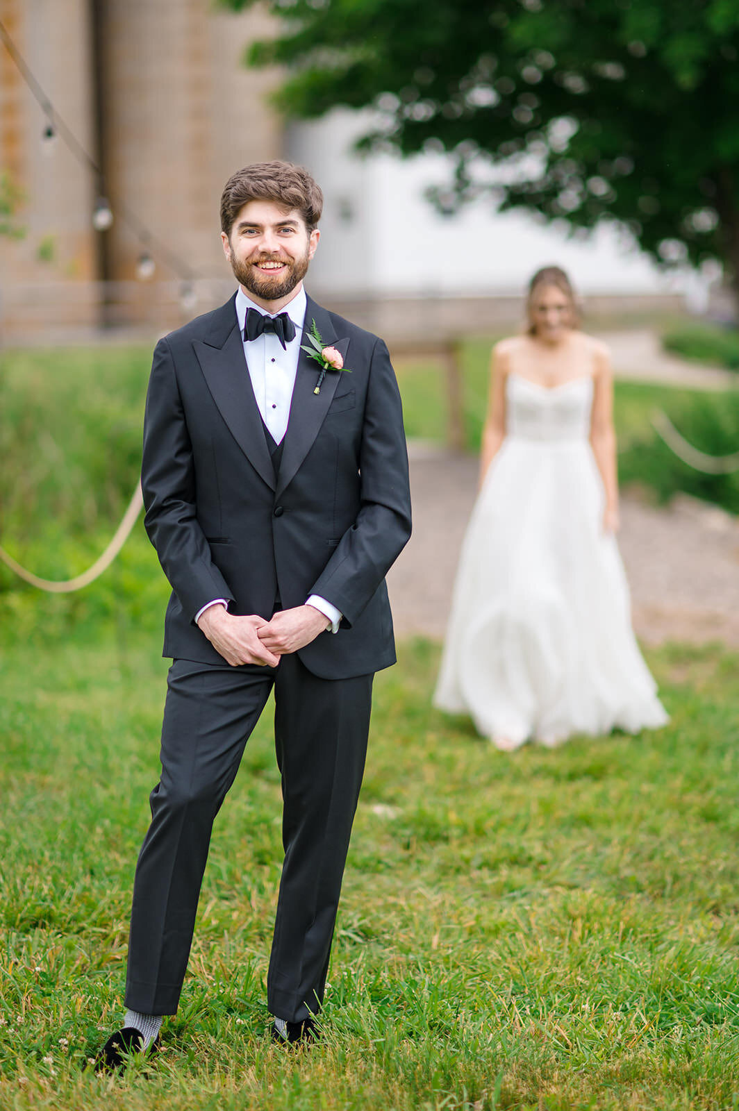 A groom in a classic black tuxedo and bow tie stands handsomely with a bride in a flowing gown blurred in the background in a pastoral setting